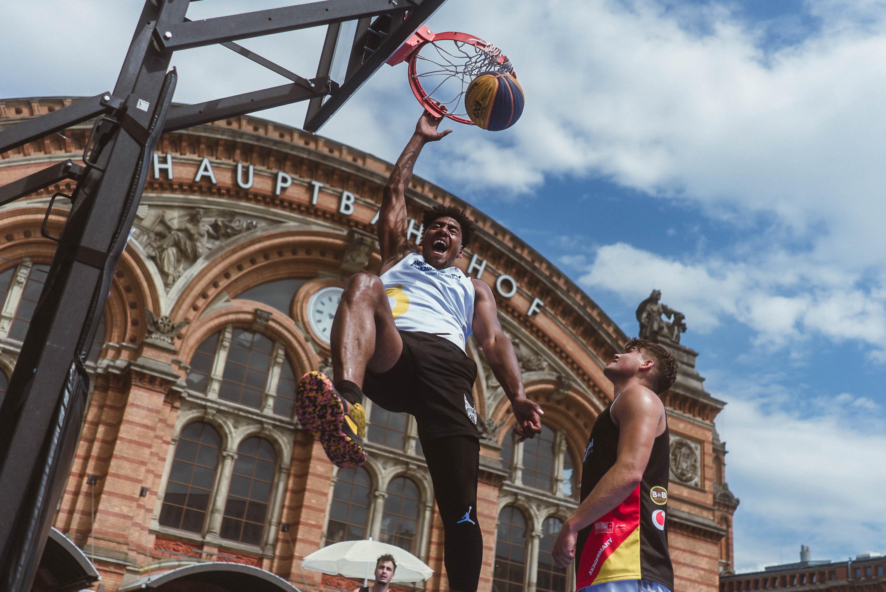 A basketball player wearing a white shirt and black trousers dunks a basketball into a basketball hoop on the forecourt of Bremen Hauptbahnhof with a cheerful expression on his face.	
