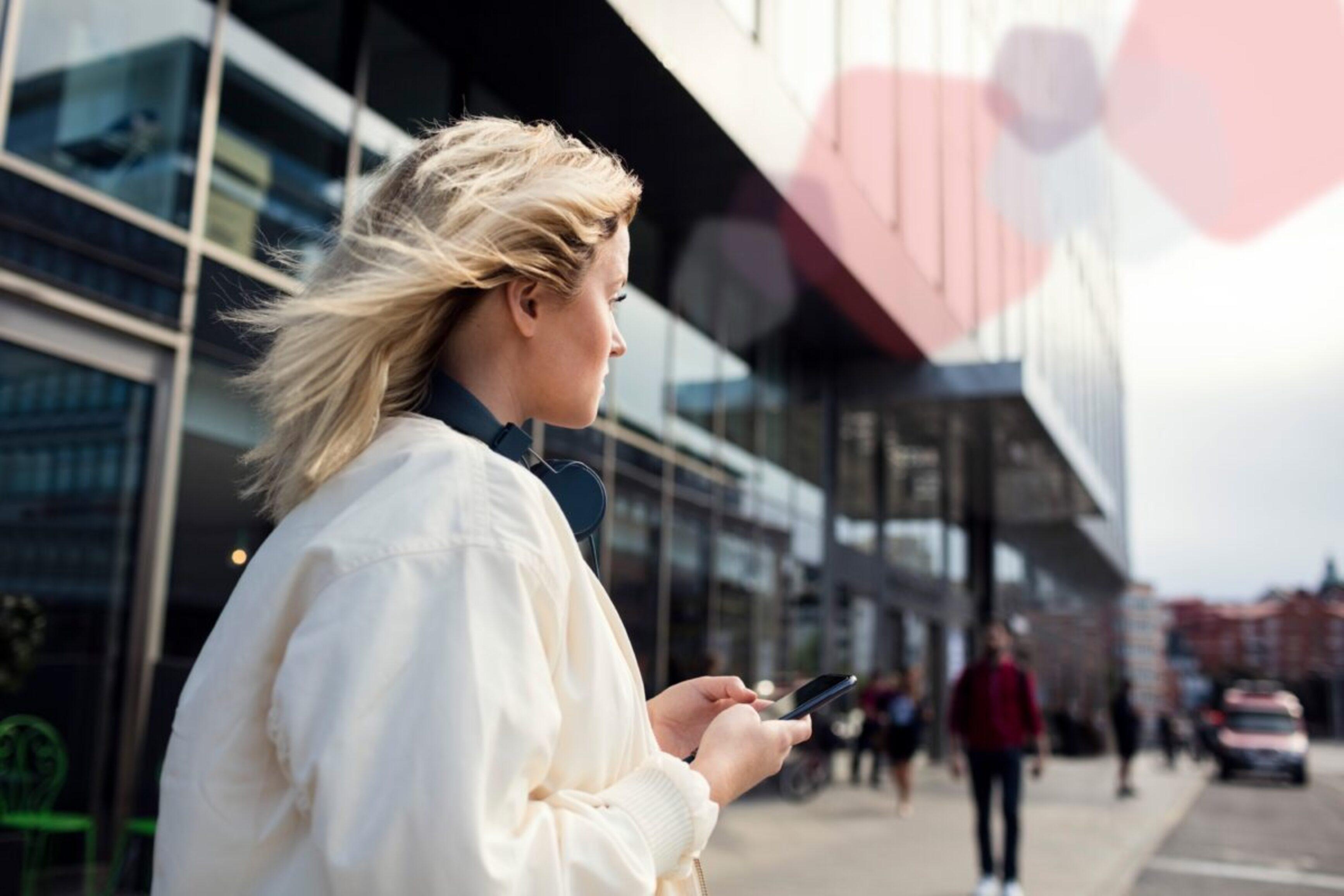 Eine Frau steht mit ihrem Smartphone vor einem Bahnhof und blickt in die Ferne.
