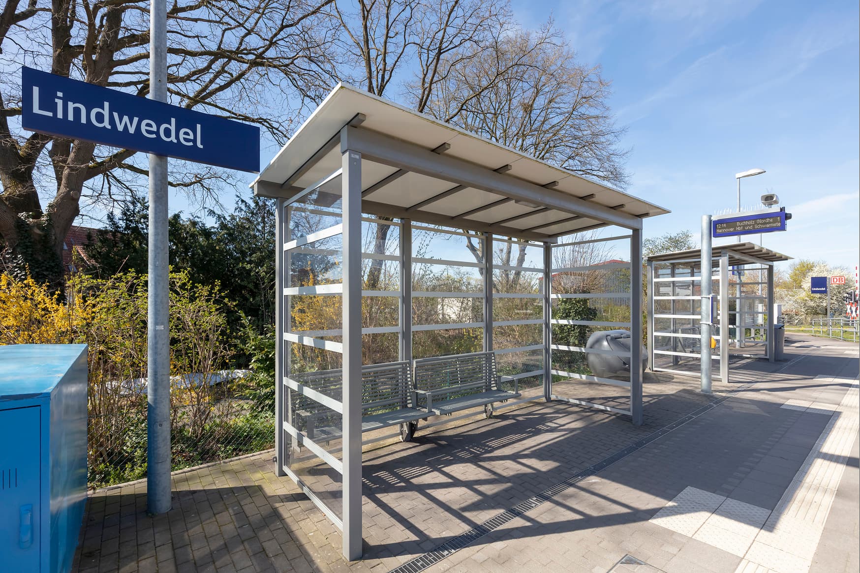 Blick auf ein Wetterschutzhäuschen auf einem Bahnsteig. Darunter eine Sitzbank. Links davon ein blaues Schild mit der Aufschrift "Lindwedel".