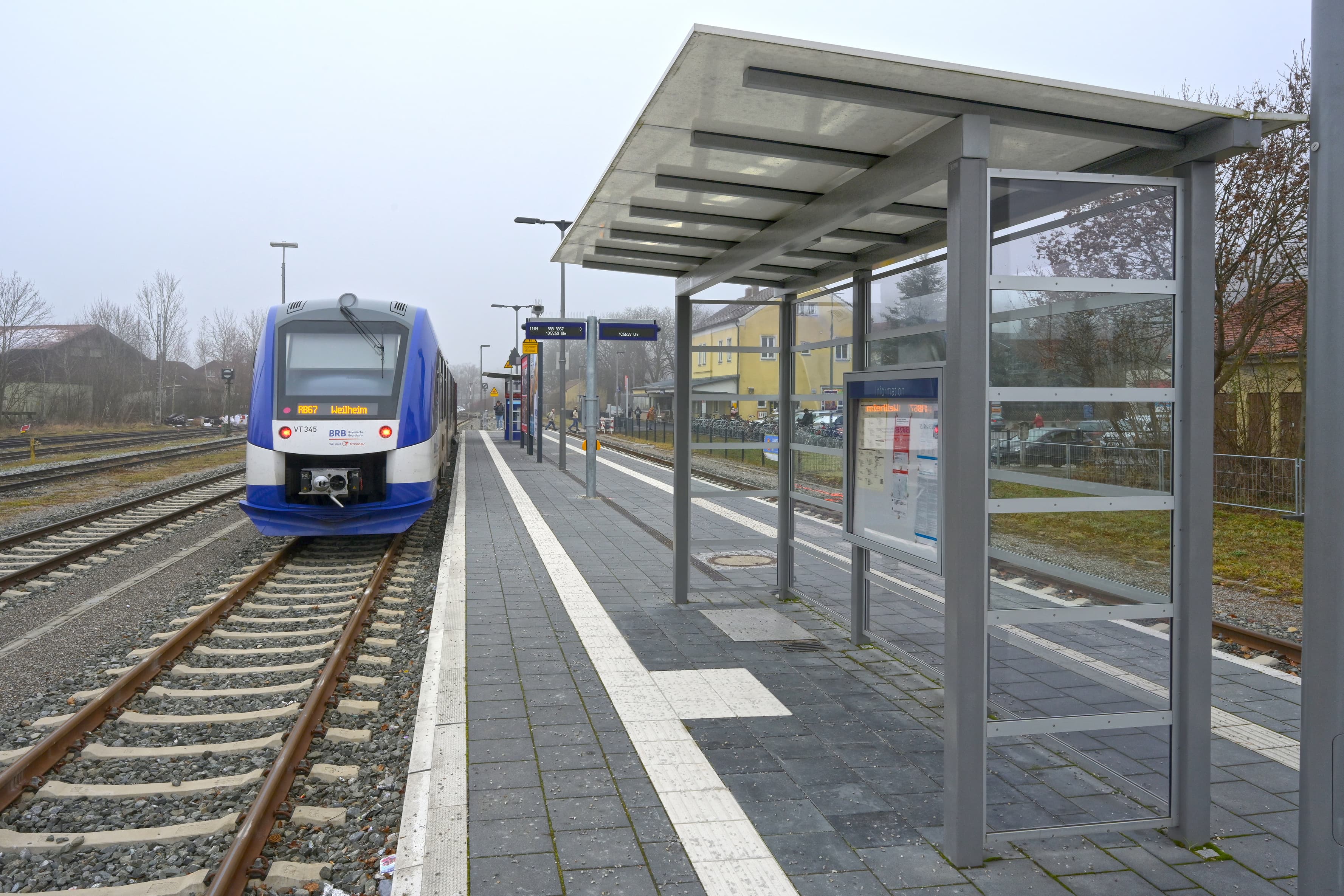 Abgebildet ein Wetterschutzhaus mit transparenten Paneelen. Dieses steht auf einem Bahnsteig. Auf der linken Seite Gleise und ein Zug. Auf der rechten Seite ebenfalls ein Gleis.