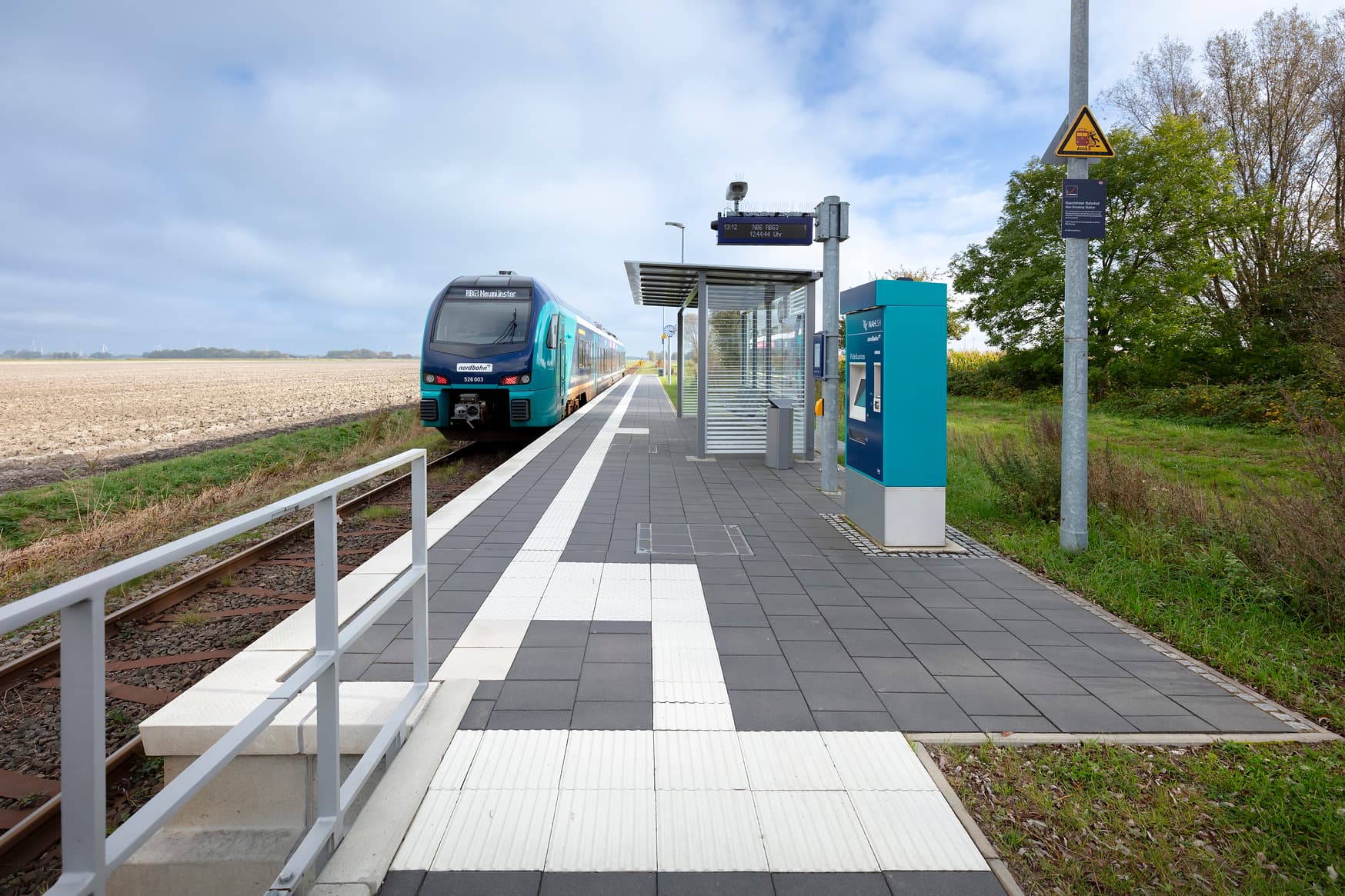Auf gepflastertem Untergrund stehen ein Wetterschutzhäuschen, ein Fahrkartenautomat und ein Zugzielanzeiger mit DAB (Digitaler Bauinformation).
