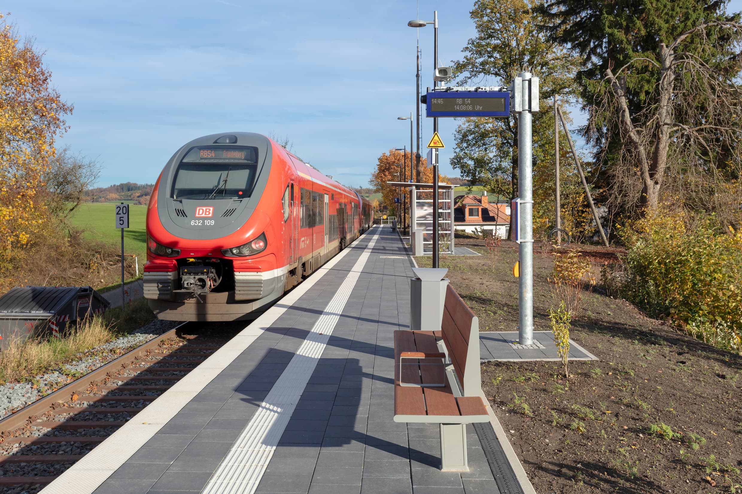 Bahnsteig mit einfahrenden Zug auf dem Gleis. Auf dem Bahnsteig ist ein taktiles Leitsystem, eine Sitzbank aus Holz, ein frisch angelegtes Beet entlang es Bahnsteigs, ein Dynamischer Schriftenanzeier und weiter hinten das Wetterschutzhäuschen.