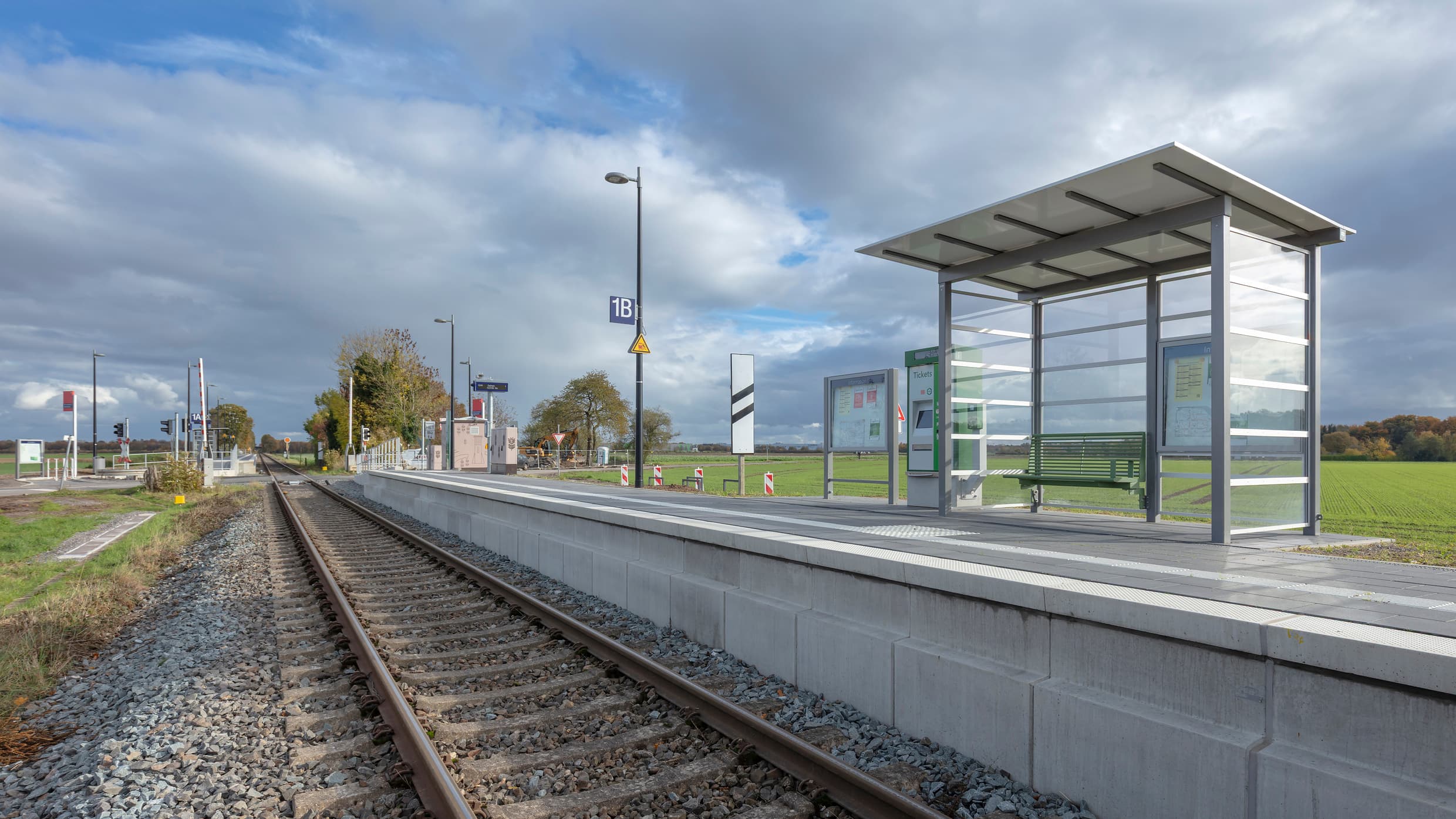 Bahnsteig mit Wetterschutzhäuschen, darin eine grüne Sitzbank. Taktiles Leitsystem auf dem Boden. Links befindet sich das Gleis. Rechts vom Bahnsteig grüne Wiese.