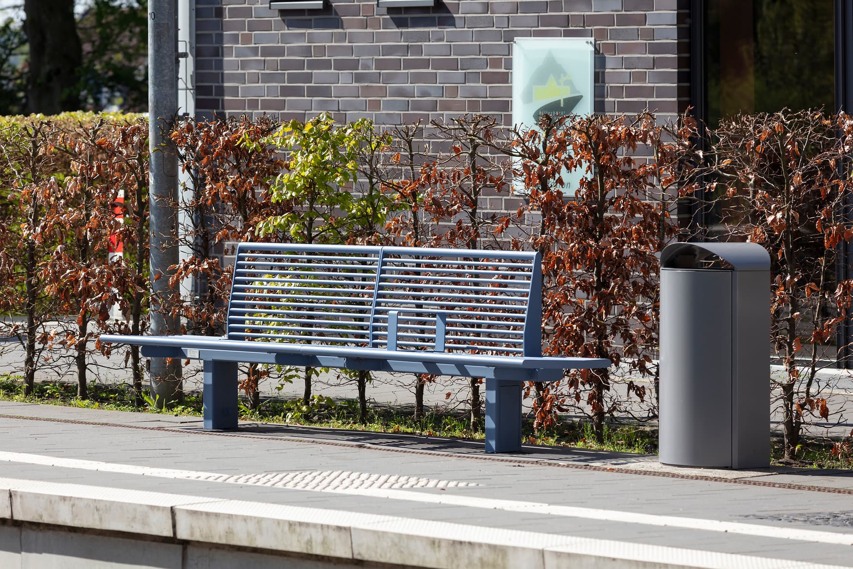 Taubenblaue Sitzbank auf Stahl auf dem Bahnsteig. Daneben eine Mülleimer.
