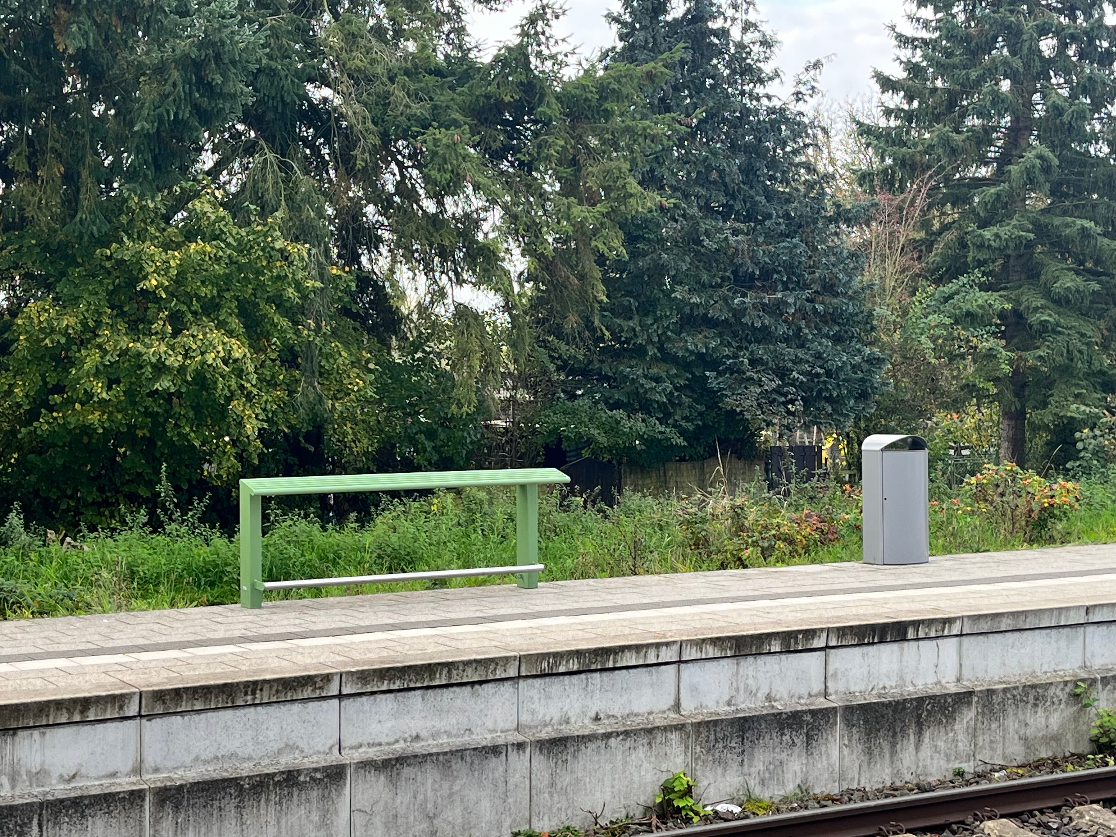 Grüner Anlehner und Metallmülleimer auf dem Bahnsteig vor grünen Bäumen. 