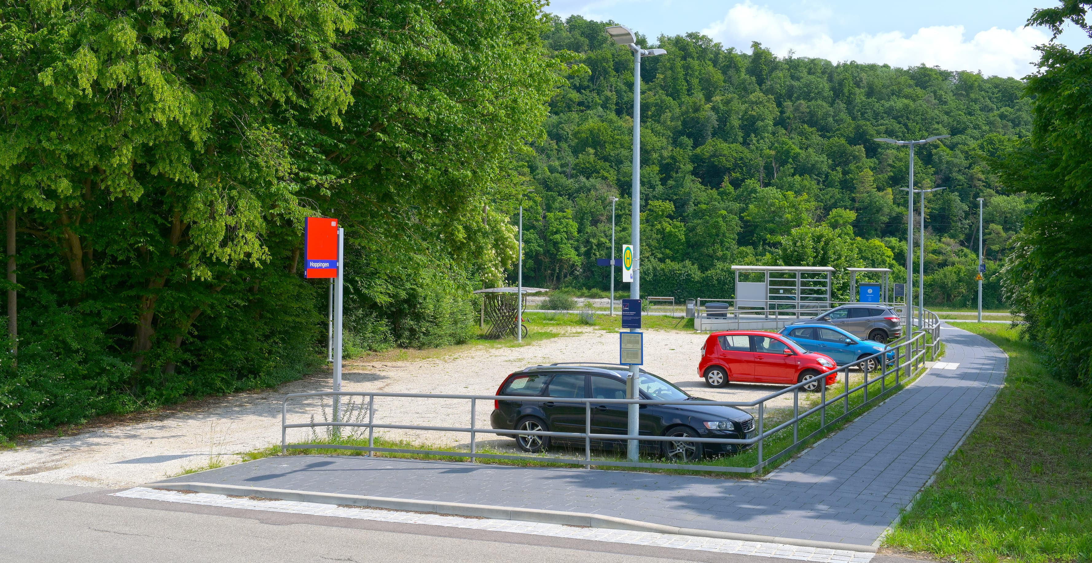 Im Vordergrund eine stufenlose Zuwegung zum Bahnsteig im Hintergrund. Links im Bild ein Schotterparkplatz, welcher direkt an den Bahnsteig grenzt. 
