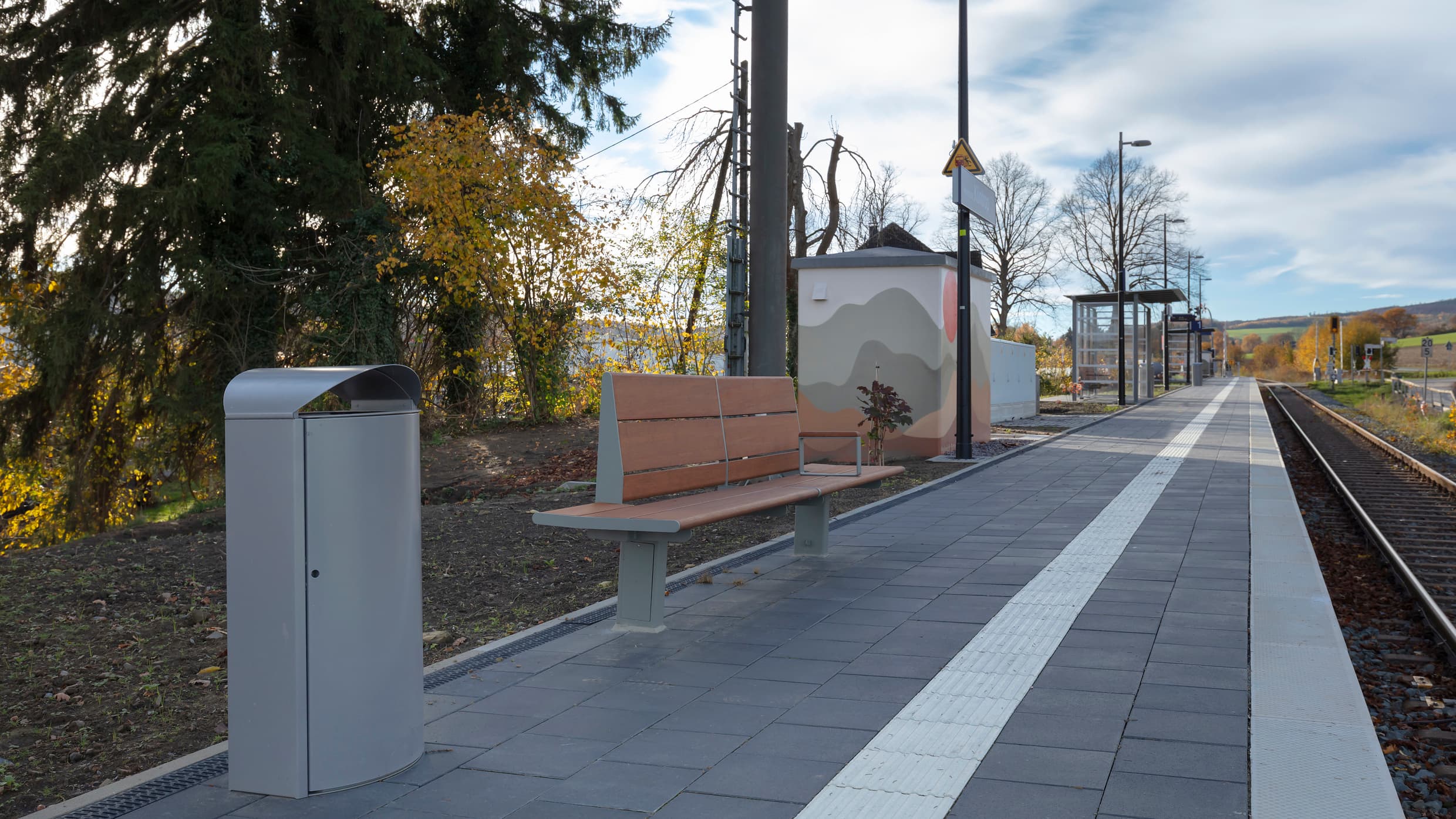 Der Bahnsteig mit taktilem Leitsystem, einem Mülleimer, einer Holzbank, den Lampen und einem bemalten Technikhäuschen.