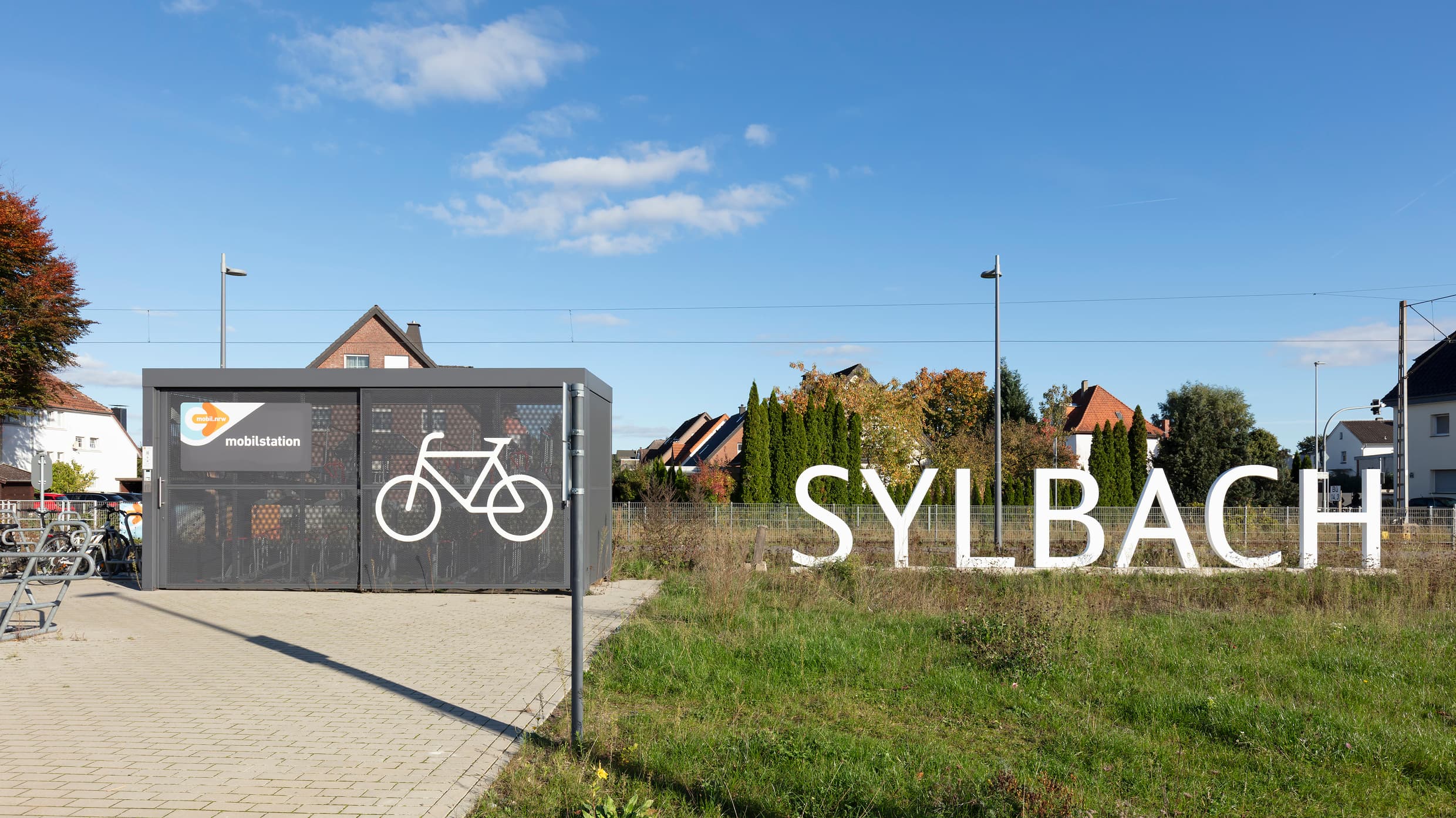 Auf einer Fahrradabstellanlage ist ein großes weißes Rad gemalt. In der Wiese daneben der personengroße Schriftzug Sylbach.