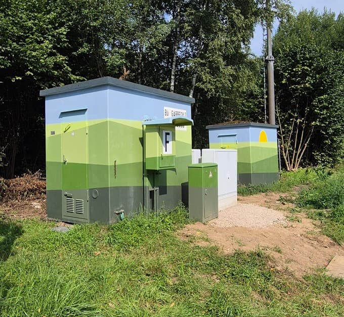 Zwei Trafohäuschen stehen auf einer Wiese vor Bäumen. Die kleinen Gebäude sind im unteren Bereich in verschiedenen Grüntönen gestrichen, im oberen Teil blau mit einer halben Sonne. Es erinnert an eine Landschaftsdarstellung.