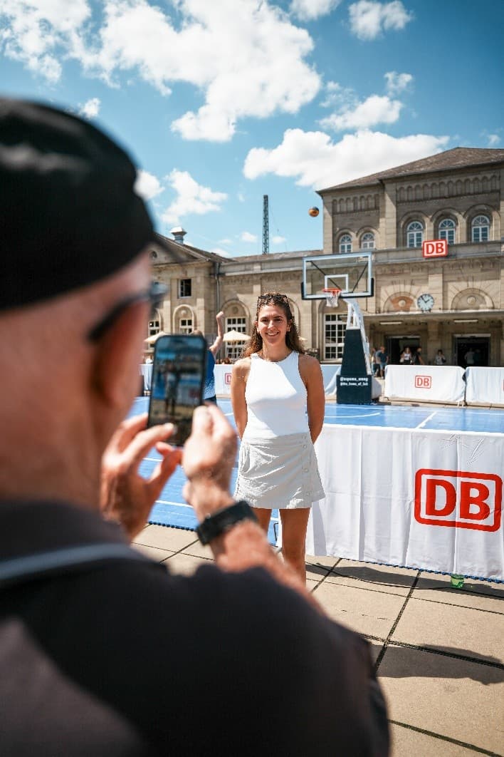 Olympiasiegerin Marie Reichert steht vor dem historischen Bahnhofsgebäude in Göttingen, im Hintergrund ist ein Basketballcourt zu sehen und unscharf im Vordergrund eine Person, die ein Foto mit dem Handy macht.