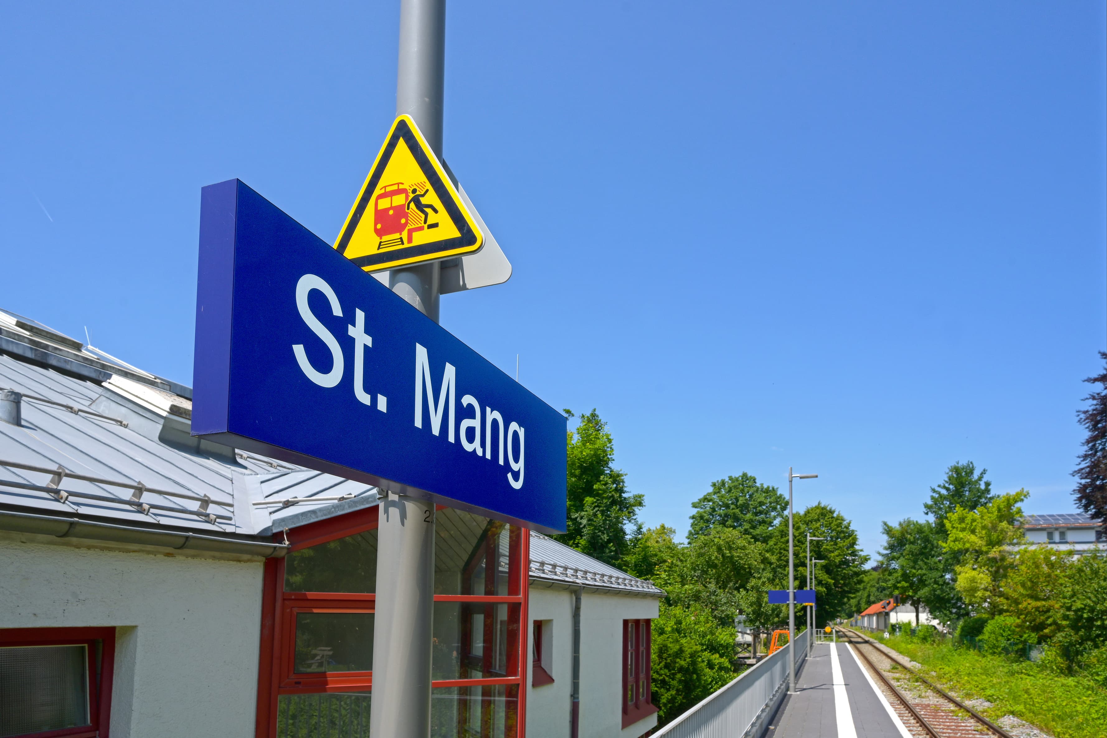Im Vordergrund das Bahnhofsschild mit dem Stationsnamen St. Mang. Im Hintergrund ein Teil das Bahnsteigs und die Gleise.