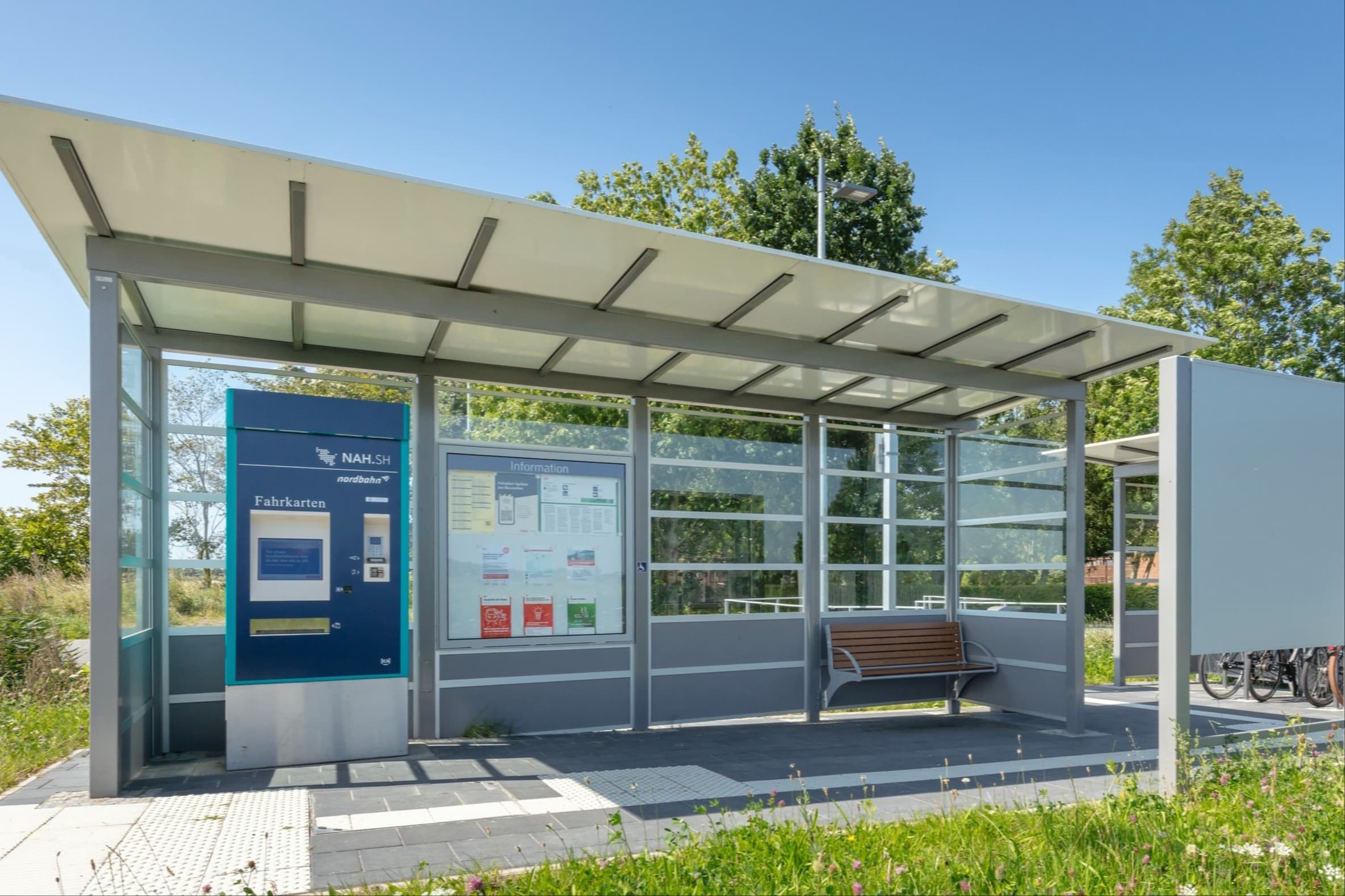 Überdachter, gläserner Wartebereich mit Fahrkartenautomat, Vitrine, Sitzbank. Im Hintergrund die Abstellmöglichkeit für Fahrräder.