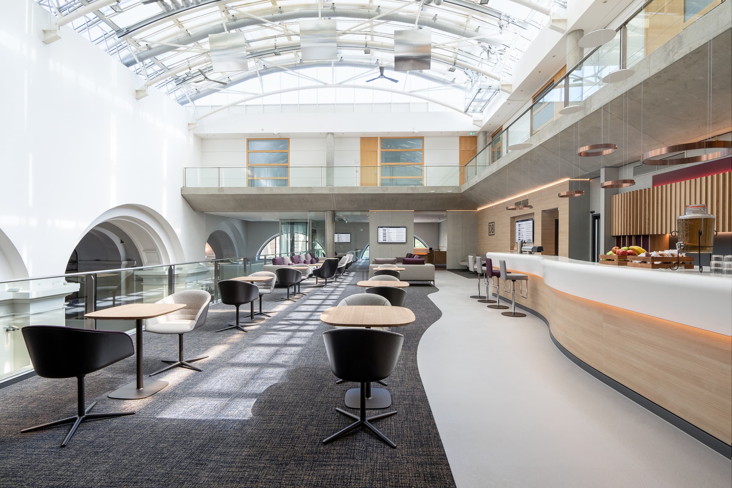 Die Innenräume der DB Lounge Frankfurt (Main) Hauptbahnhof mit Sitzgelegenheiten und einer Servicetheke, überdacht von einem bogenförmigen Glasdach.