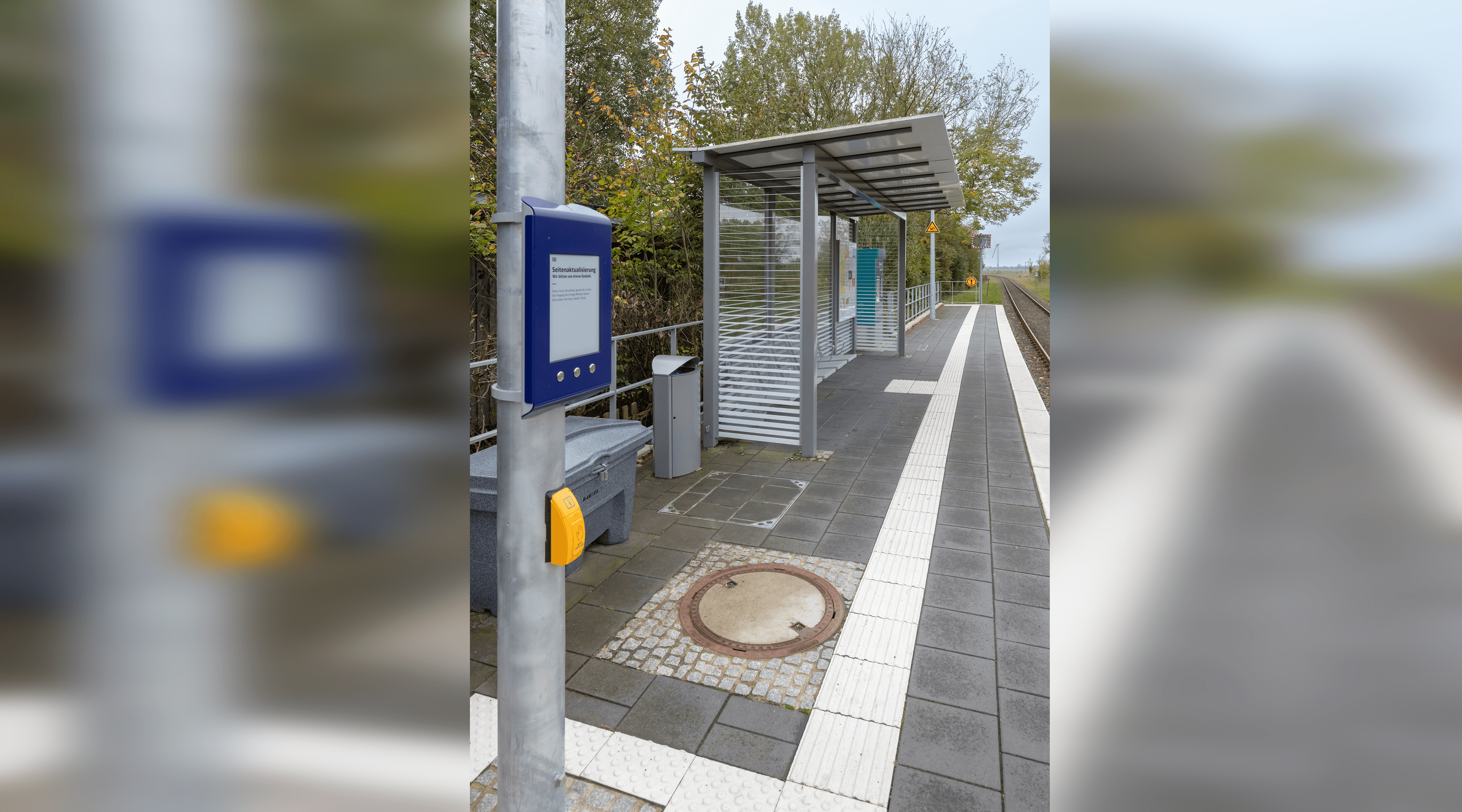 Digitaler Aushang Bau Kommunikation (DAB) mit gelber Taste zum Vorlesen an einer Stele auf dem Bahnsteig. Daneben das Wetterschutzhäuschen.