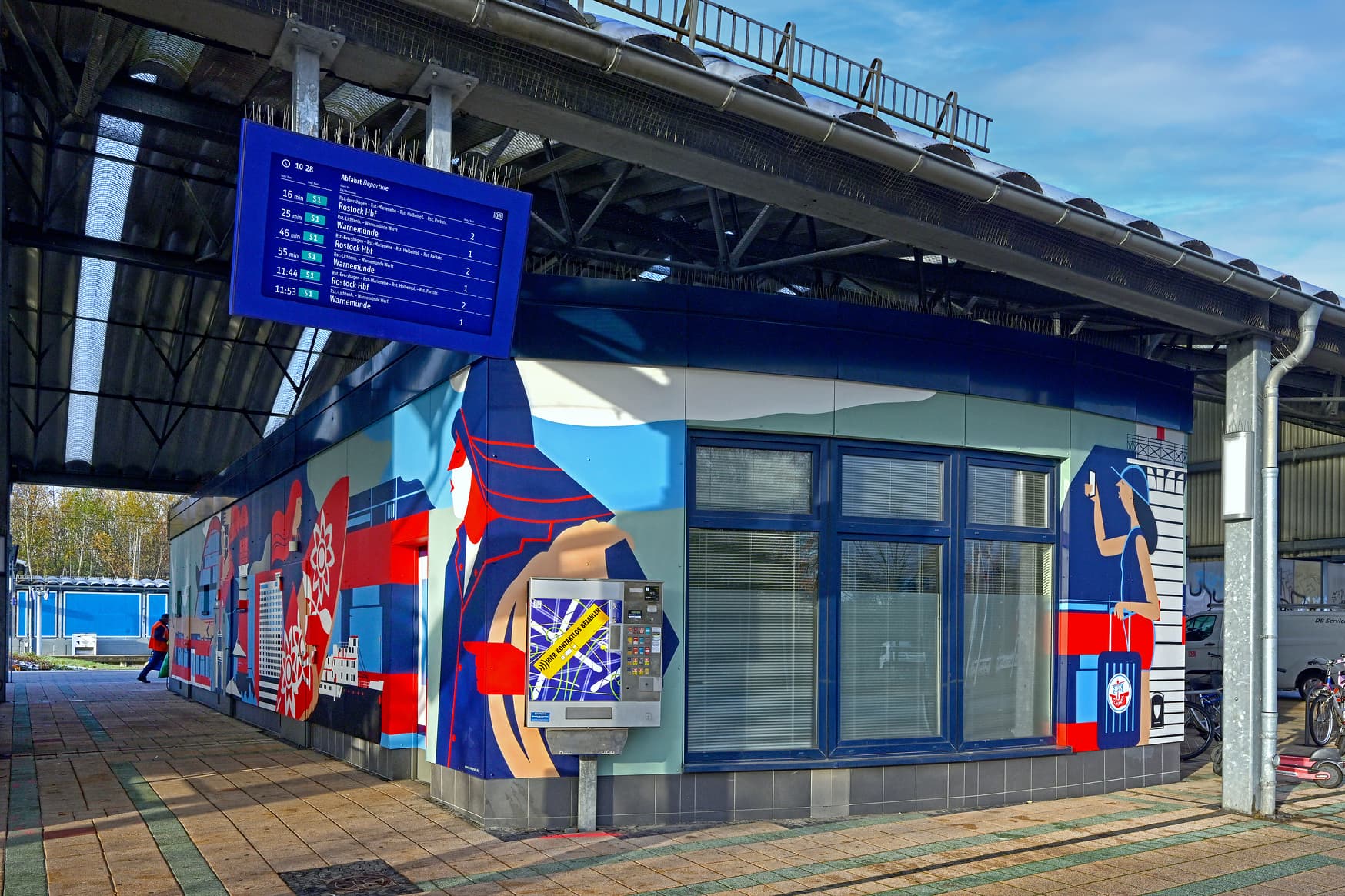 ZugInfoMonitor am Bahnsteigdach. Auf der Wand die neuen, bunten Motive vom Künstler.