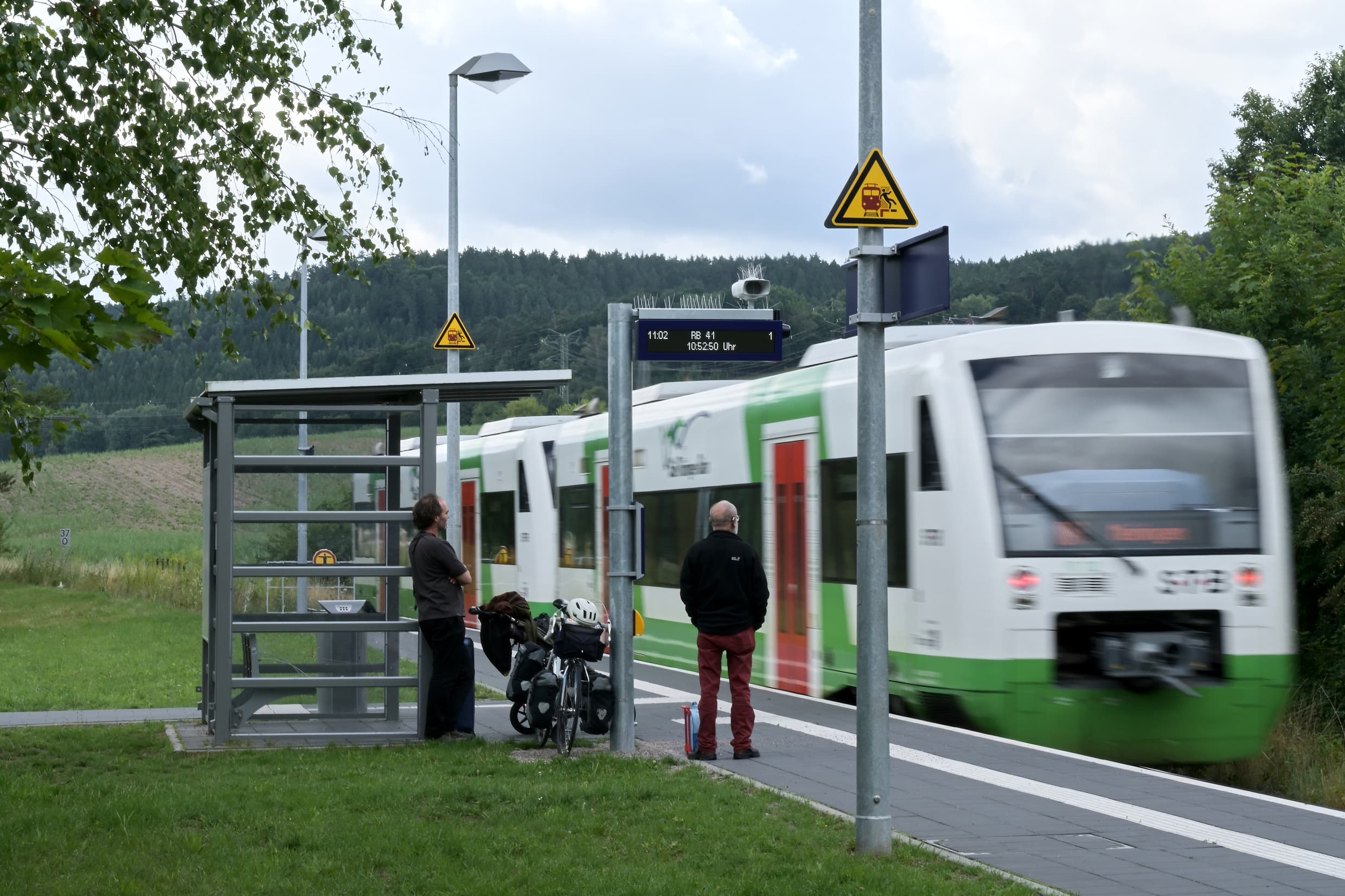 Ein einfahrender Zug am neuen Bahnsteig. Die Reisenden warten am volltransparenten Wetterschutzhaus für den Einstieg.