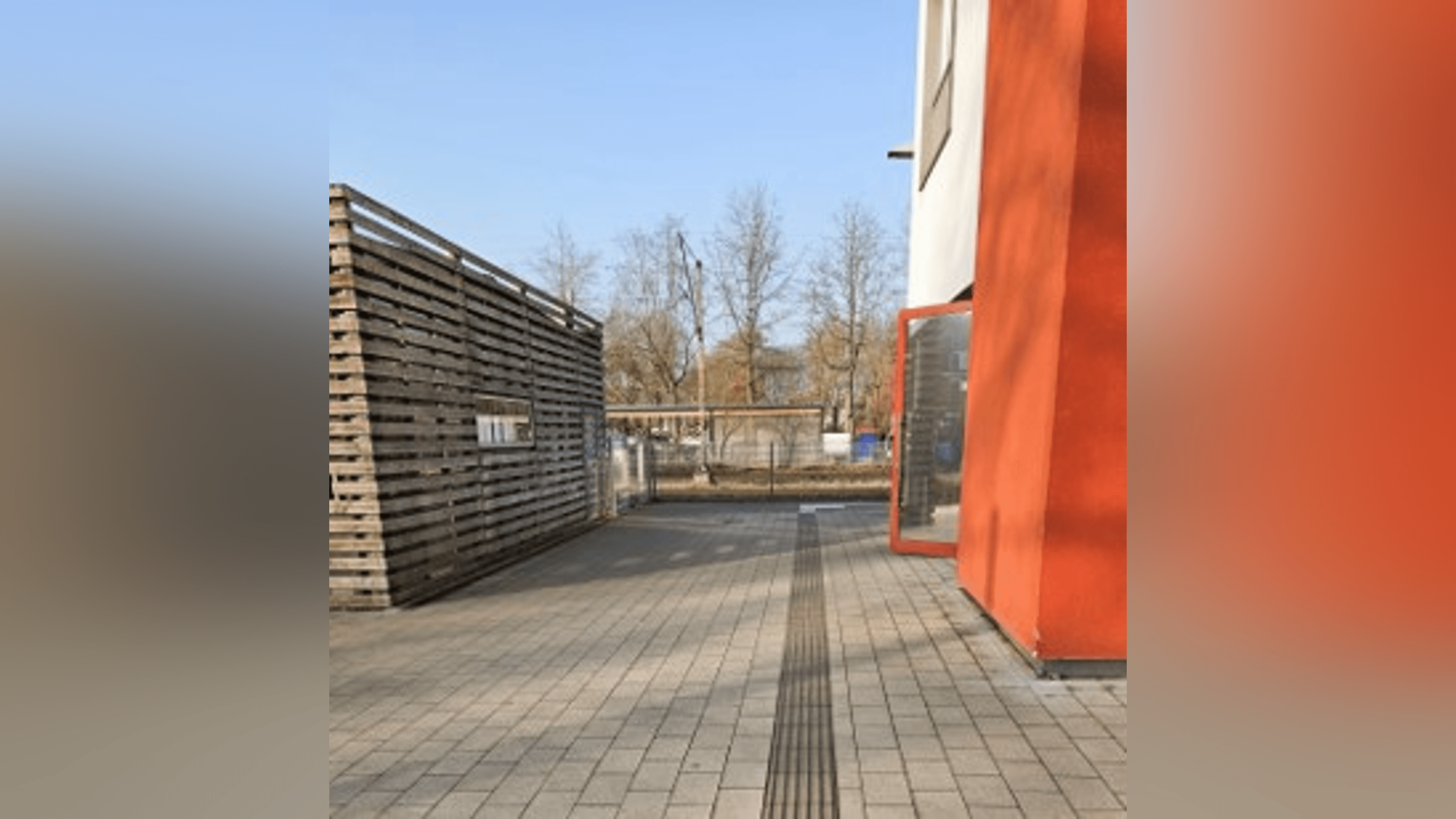 Zugang zum Bahnsteig mit Blindenleitsystem auf dem Boden, recht ein orange gestrichenes Gebäude, links ein Gebäude mit Holzfassade.