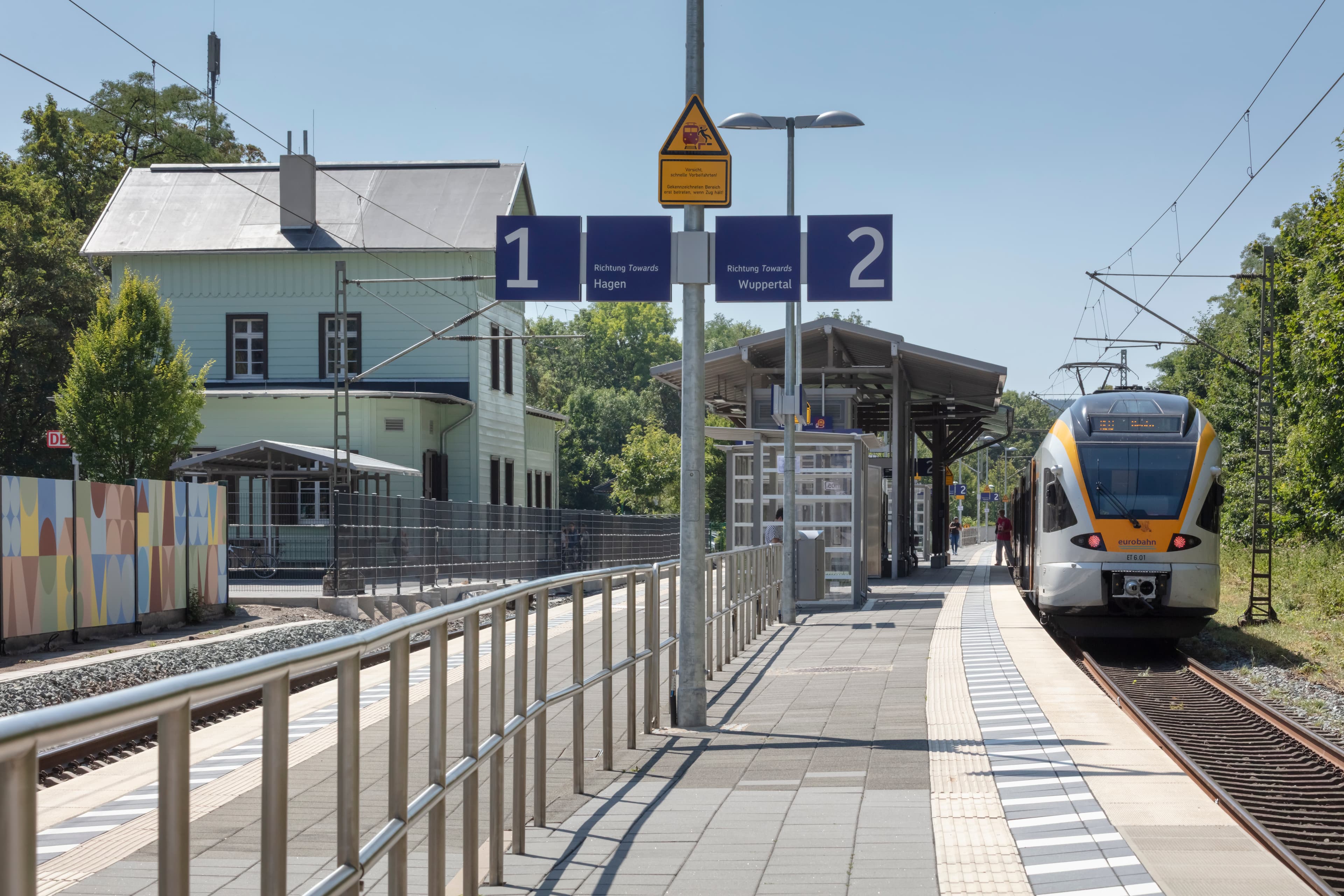 Bahnsteig mit moderner Beschilderung, farbenfrohem Wetterschutzhaus und historischem Bahnsteigdach.