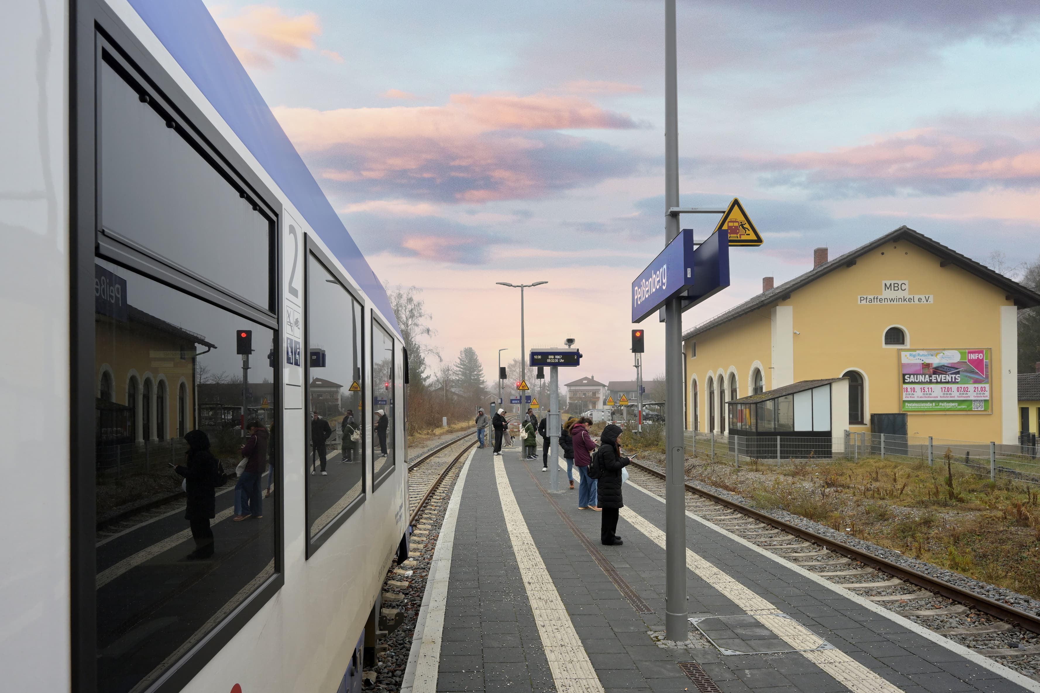 Abgebildet das Bahnhofsschild mit dem Bahnhofsnamen Peißenberg. Darunter ein moderner Bahnsteig. In der Mitte ein Mast, an dem digitale Anzeigegeräte befestigt sind. Im Hintergrund sind Menschen auf dem Bahnsteig. Links ein Zug auf einem Gleis. Rechts ebenfalls ein Gleis. Rechts ein Gebäude.