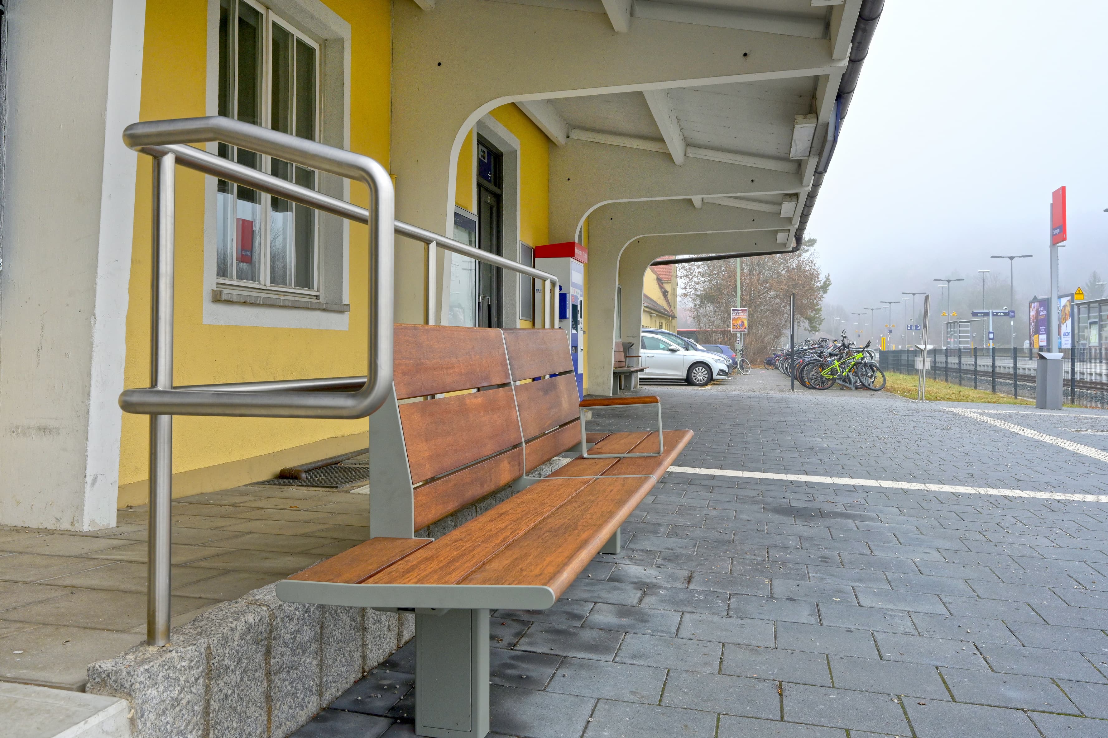Abgebildet eine Sitzbank mit Holzbelag. Links davon ein Gebäude. Rechts im Hintergrund ein Bahnsteig.