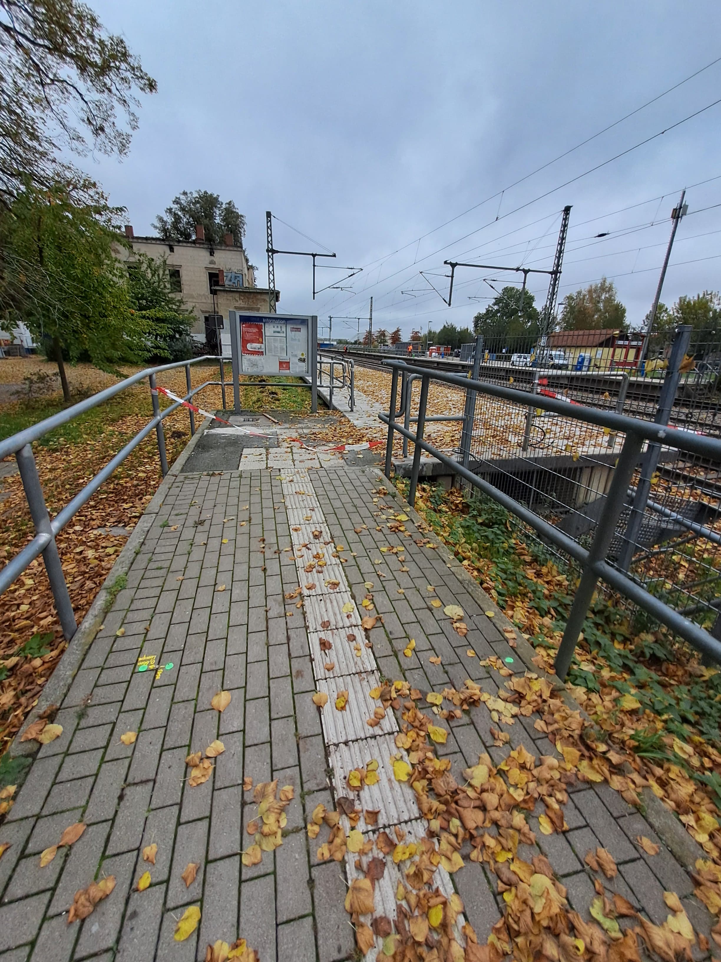 Ein gepflasterter Weg mit Geländer und Laub mit rudimentärem Blindenleitstreifen, rechts ein Bahnsteig.