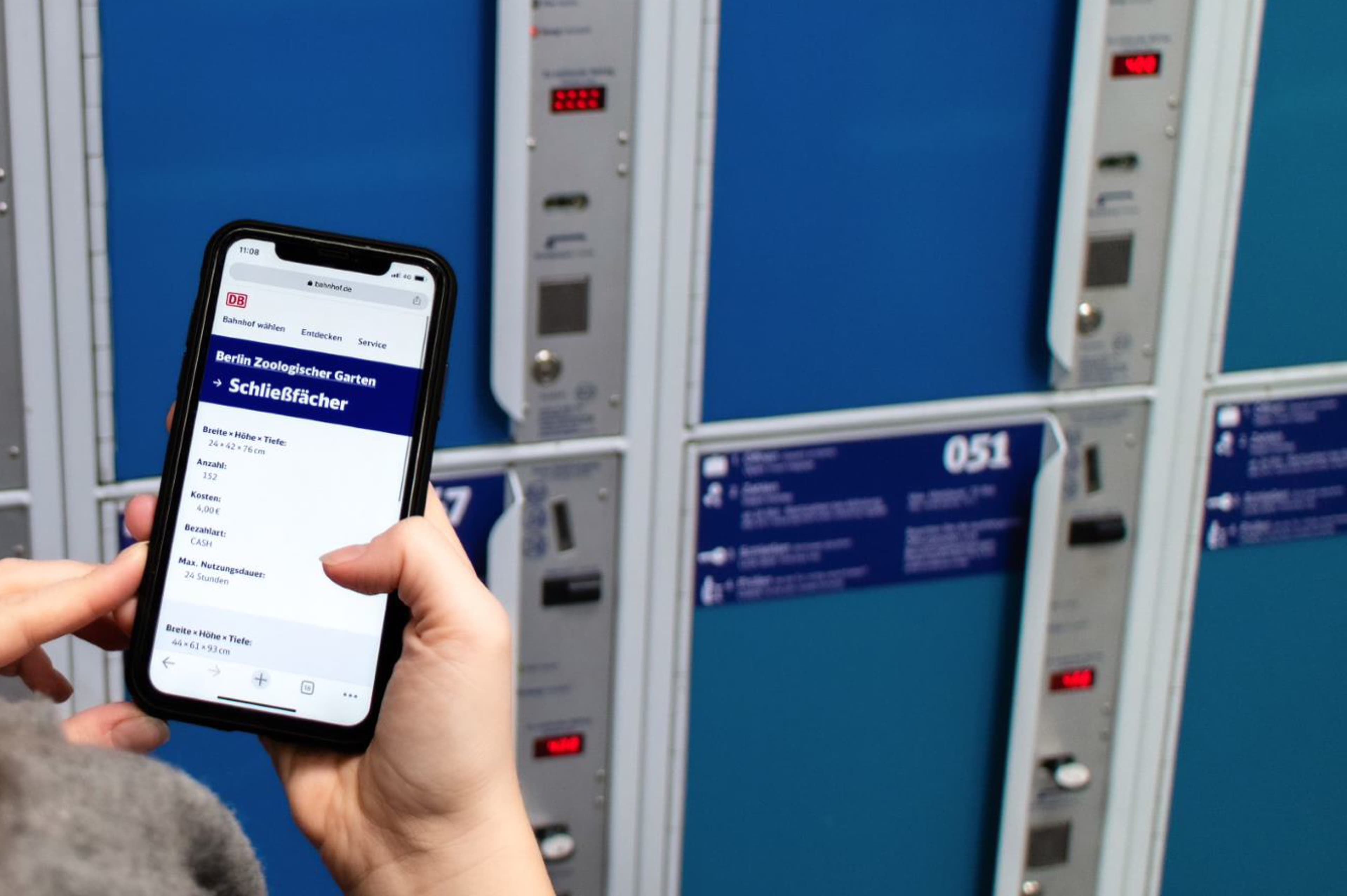 Eine Person steht vor den Gepäckschließfächern eines Bahnhofs und hat eine entsprechende Seite von bahnhof.de auf Ihrem Smartphone geöffnet.