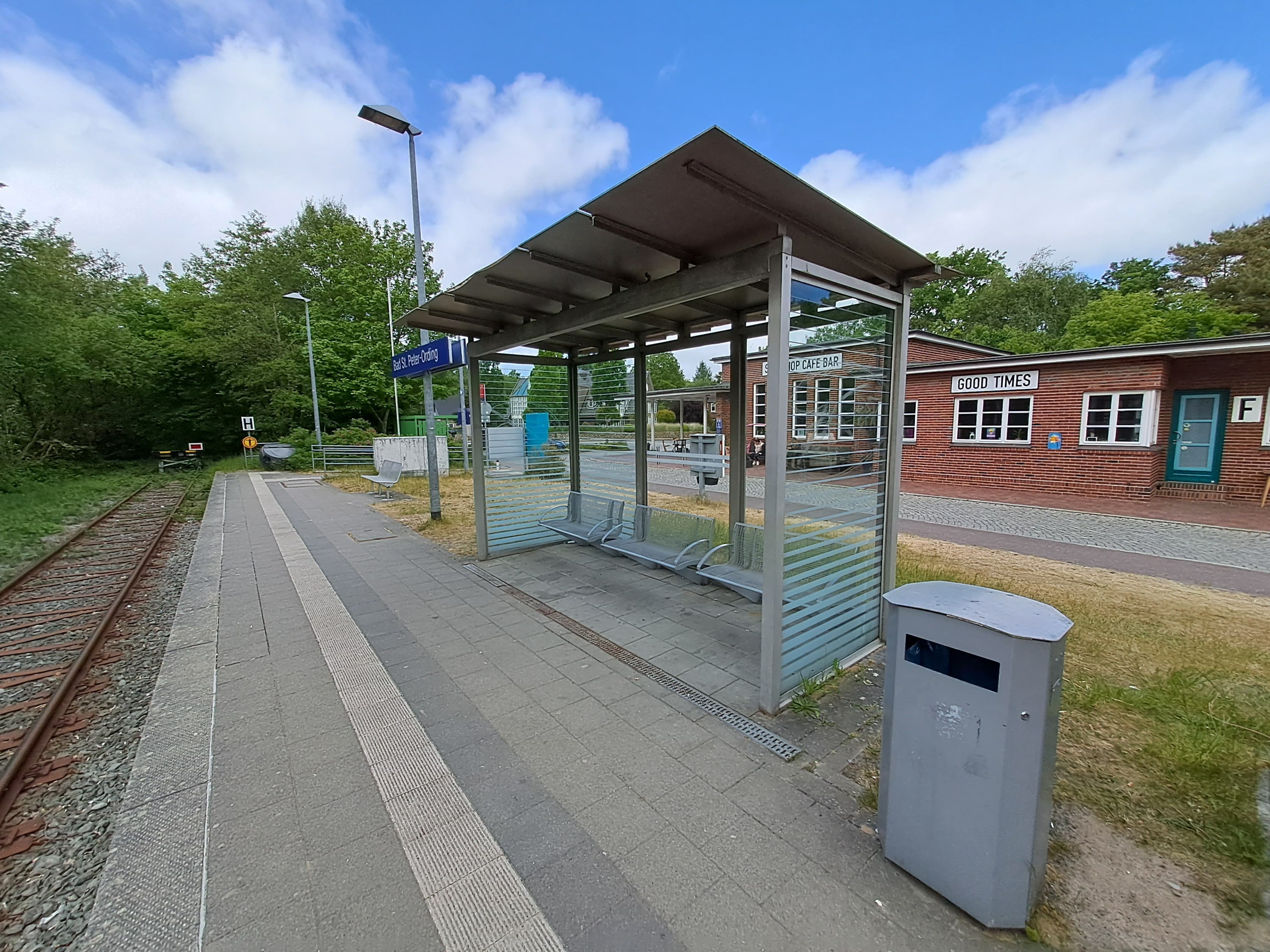 Blick auf das alte Wetterschutzhäuschen. Rechts davon ein Mülleimer. Im Hintergrund befindet sich ein gemauertes Gebäude.