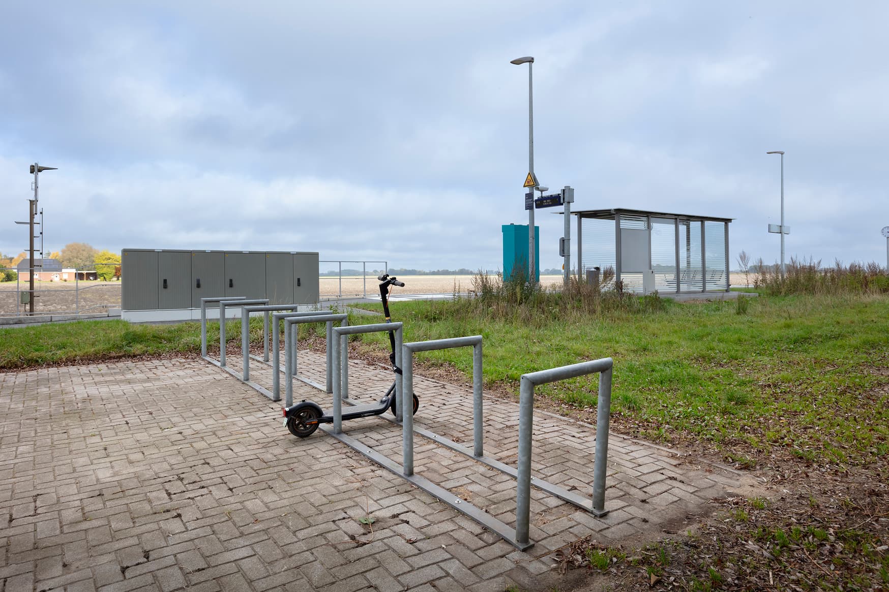 Fahrradabstellanlage am Bahnsteigzugang mit Fahrradbügeln, daran ist ein Elektroroller abgeschlossen. Im Hintergrund Stromkasten und Bahnsteig mit Wetterschutzhäuschen.