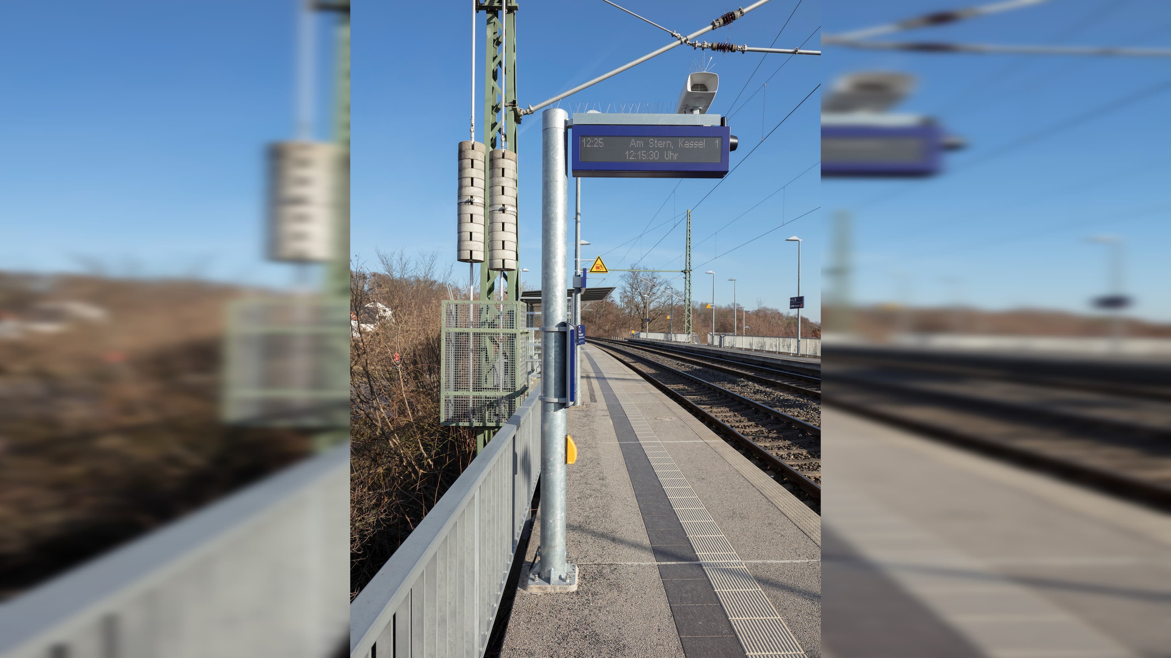 Blick auf das DSA+ Fahrgastinformationssystem. Rechts befindet sich das Gleis. Auf dem Bahnsteig ist auch ein Wetterschutzhäuschen umringt von einem Metallzaun zur Absicherung zu sehen.