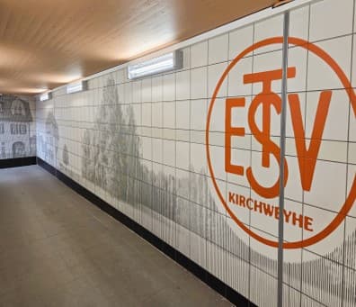 Eine Wand der Personenunterführung mit Baummotiv und im Vordergrund das orangene Logo vom ESTV Kirchweyhe.