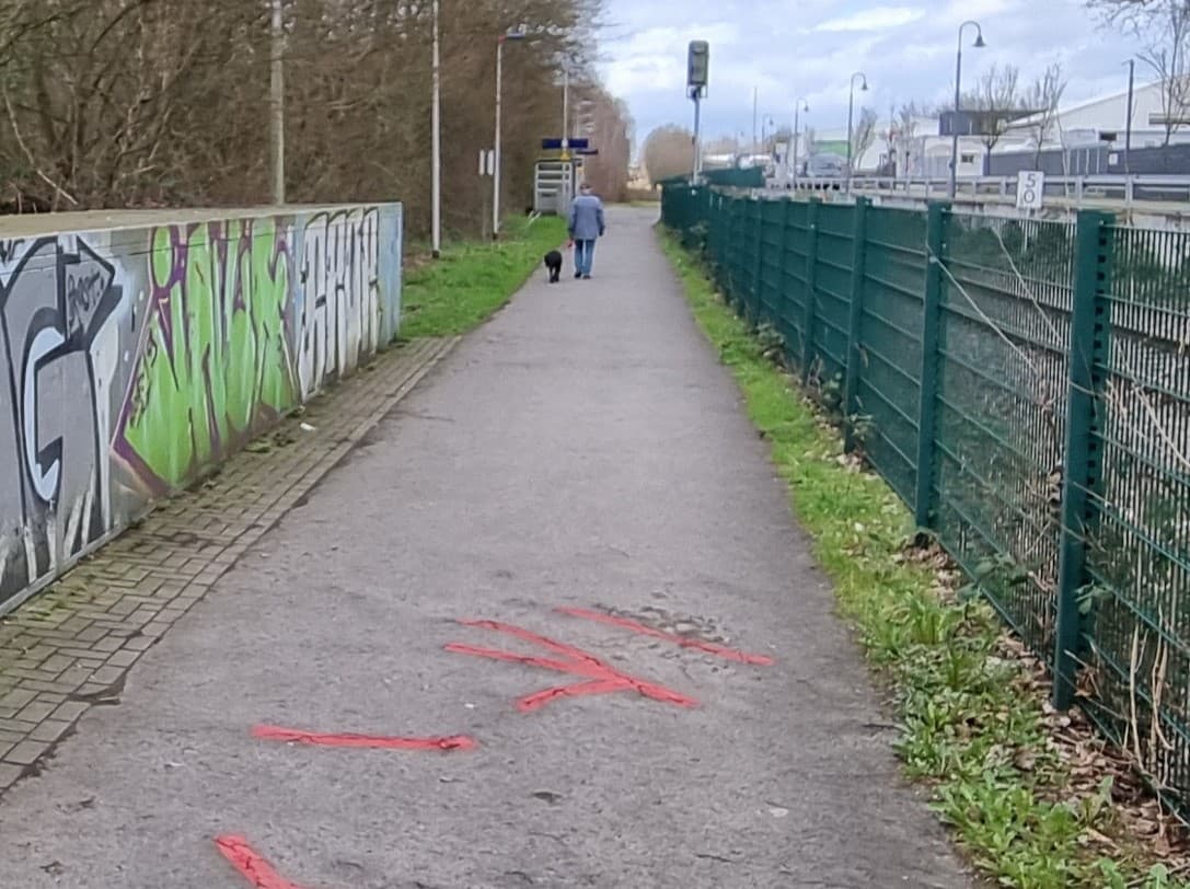Zuwegung ohne taktiles Leitsystem. Rechts ein grüner Zaun, der den Weg vom Gleis trennt. Links die Fahrradboxen mit Graffiti.