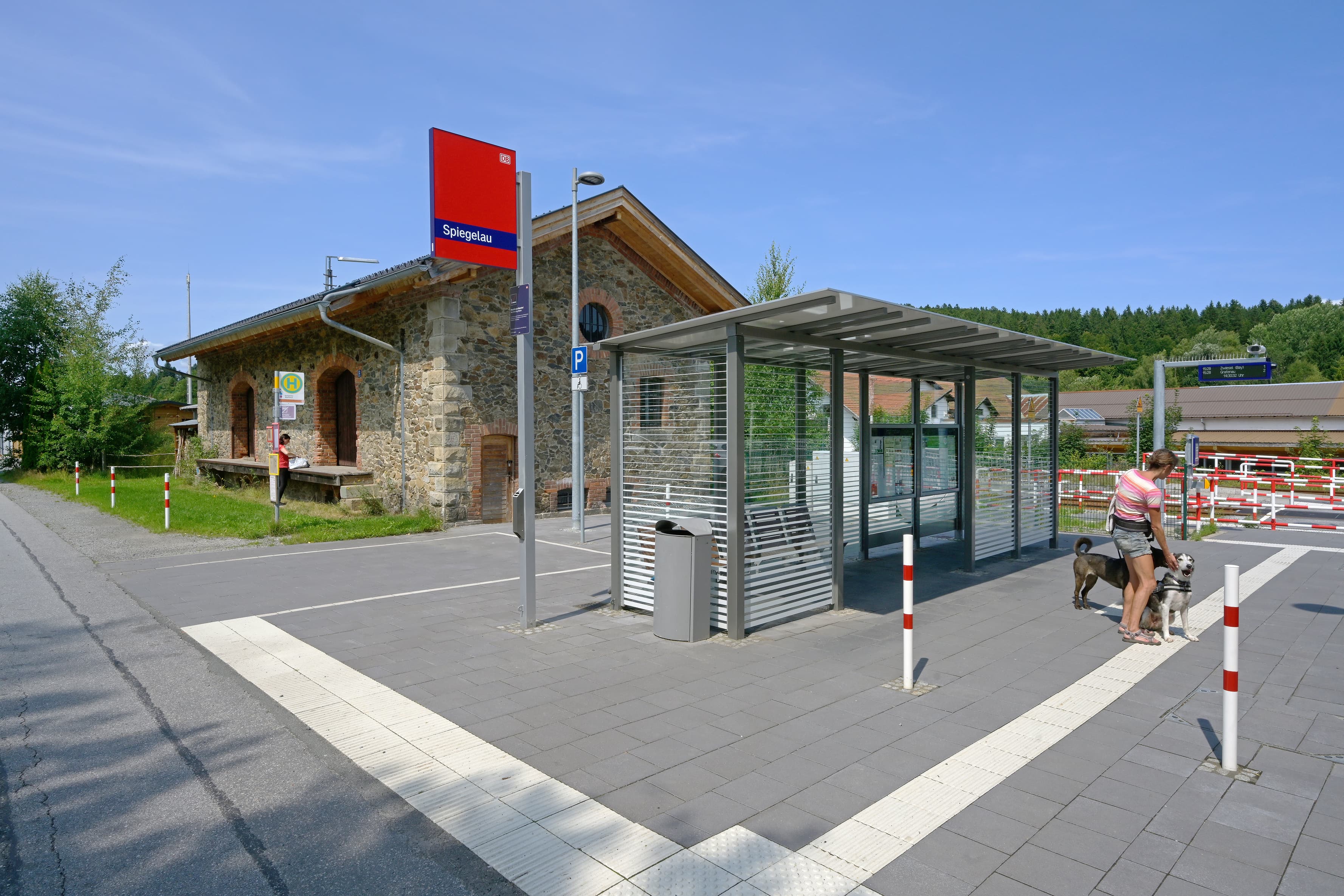 Vorne im Bild ein Wetterschutzhaus am Bahnsteig. Dahinter das alte, steinerne Bahnhofsgebäude. 