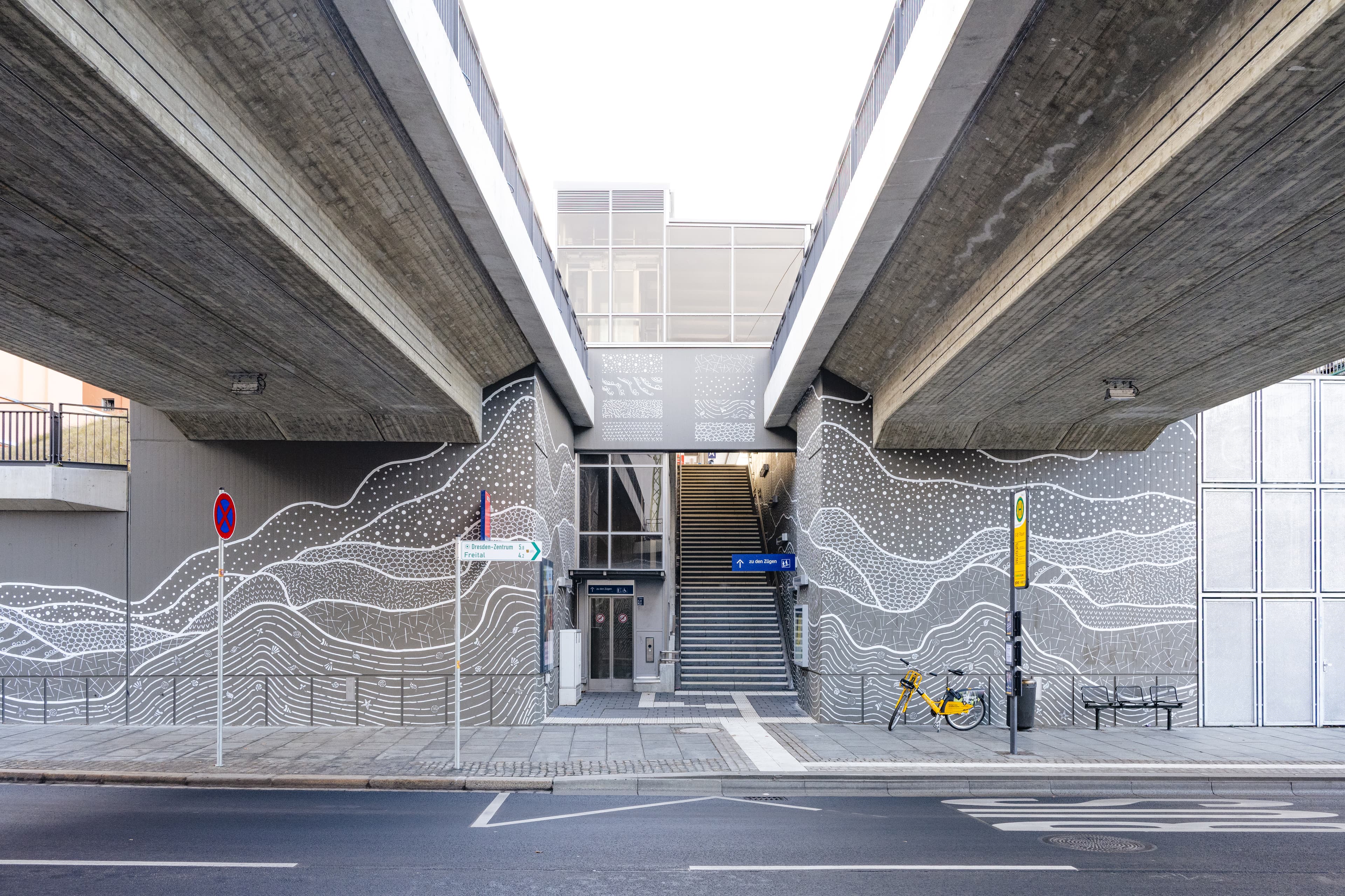 Blick auf den Zugang der Verkehrsstation: Eine Treppe führt nach oben auf den höher gelegenen Bahnsteig. Die Wände rechts und links neben dem Zugang und der Treppenbereich an sich, wurden mit einer reliefartigten Gestaltung in grau/weiß aufgewertet.