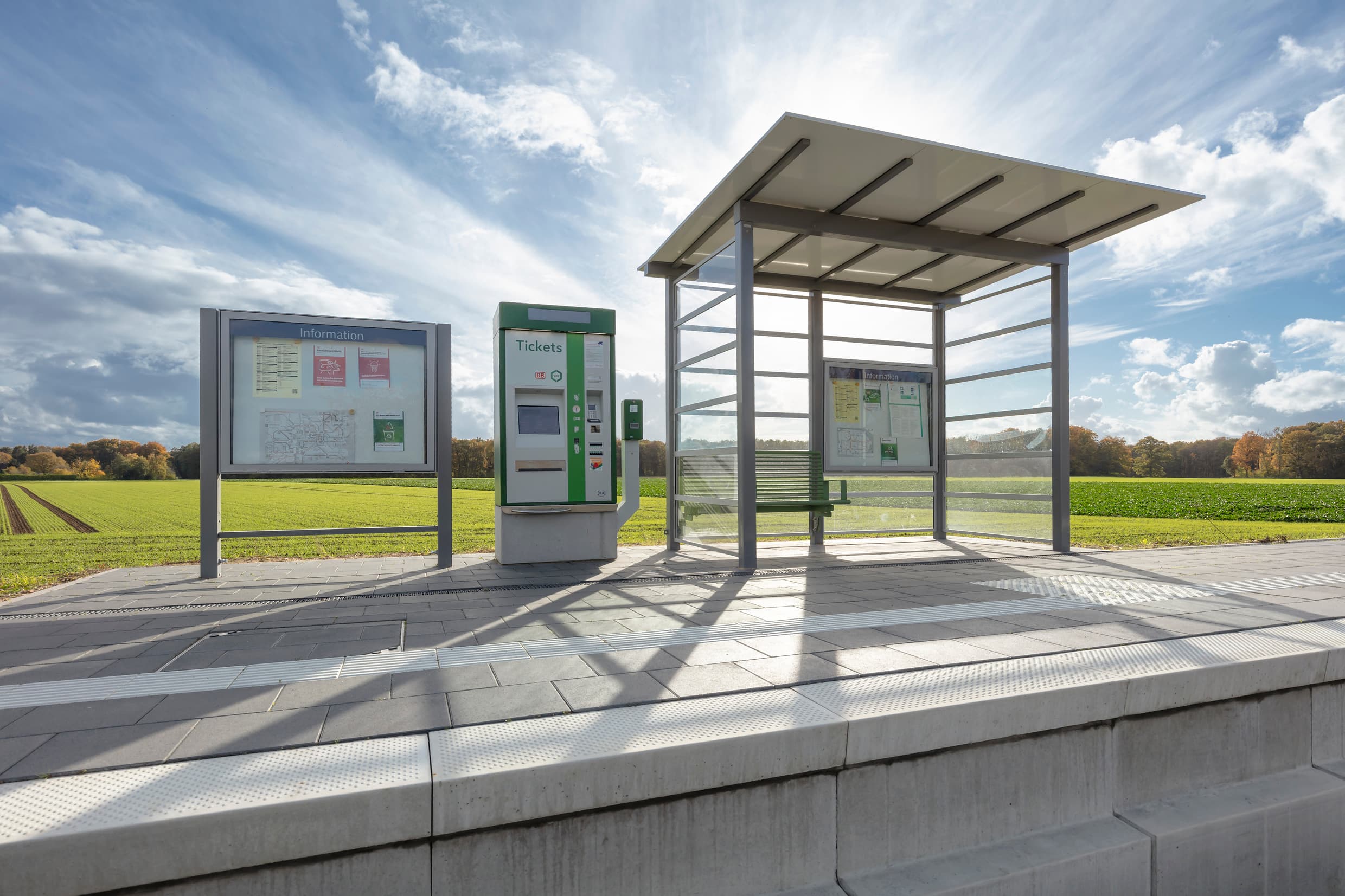 Blick auf neue Vitrine, Ticketautomat und Wetterschutzhäuschen auf dem Bahnsteig. Dahinter grüne Wiesen.