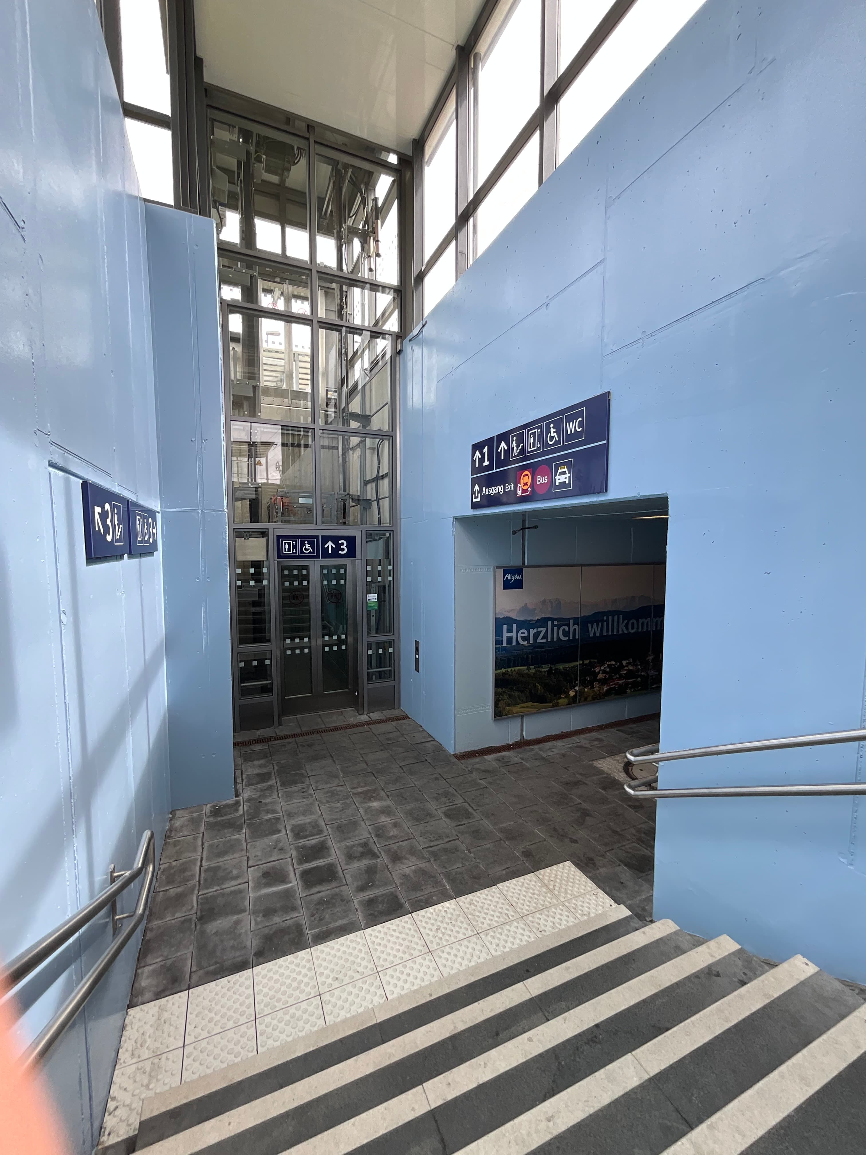 Blick von einem Bahnsteig hinab in die frisch hellblau gestrichene Personenunterführung des Bahnhofs Leutkirch im Allgäu. Im Vordergrund eine Treppe mit weißen, taktil markierten Stufen. Im unteren Bereich der Personenunterführung links ein Aufzug aus Glas und Stahl, an den Wänden blaue Wegeleitschilder zu den Bahnsteigen 1 und 3 sowie zu Aufzug, WC und Ausgang. Rechts ein großformatiges Wandbild mit Allgäuer Landschaftsmotiv und der Aufschrift „Herzlich willkommen".