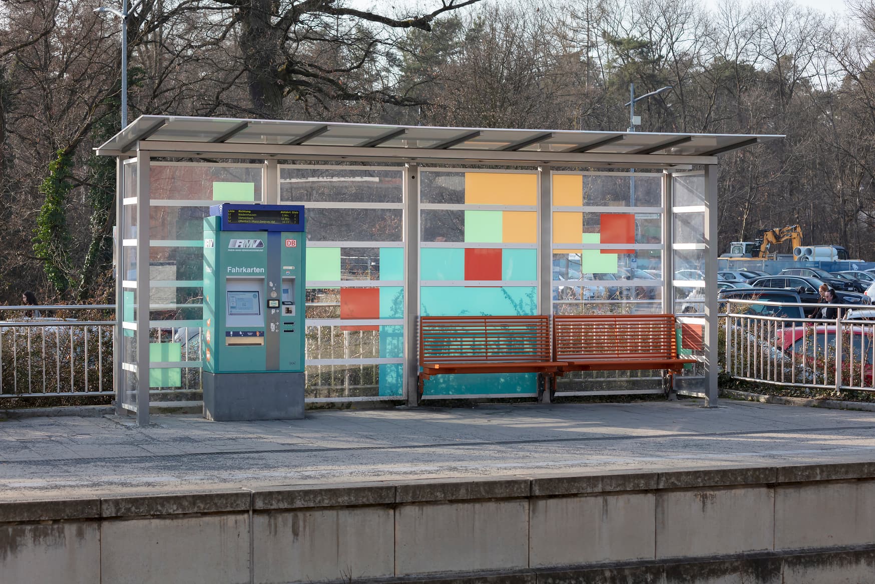 Blick auf ein Wetterschutzhäuschen. Darunter befindet sich ein Ticketautomat und rechts davon eine orange farbene Sitzbank. Im Hintergrund sind geparkte Autos zu sehen.