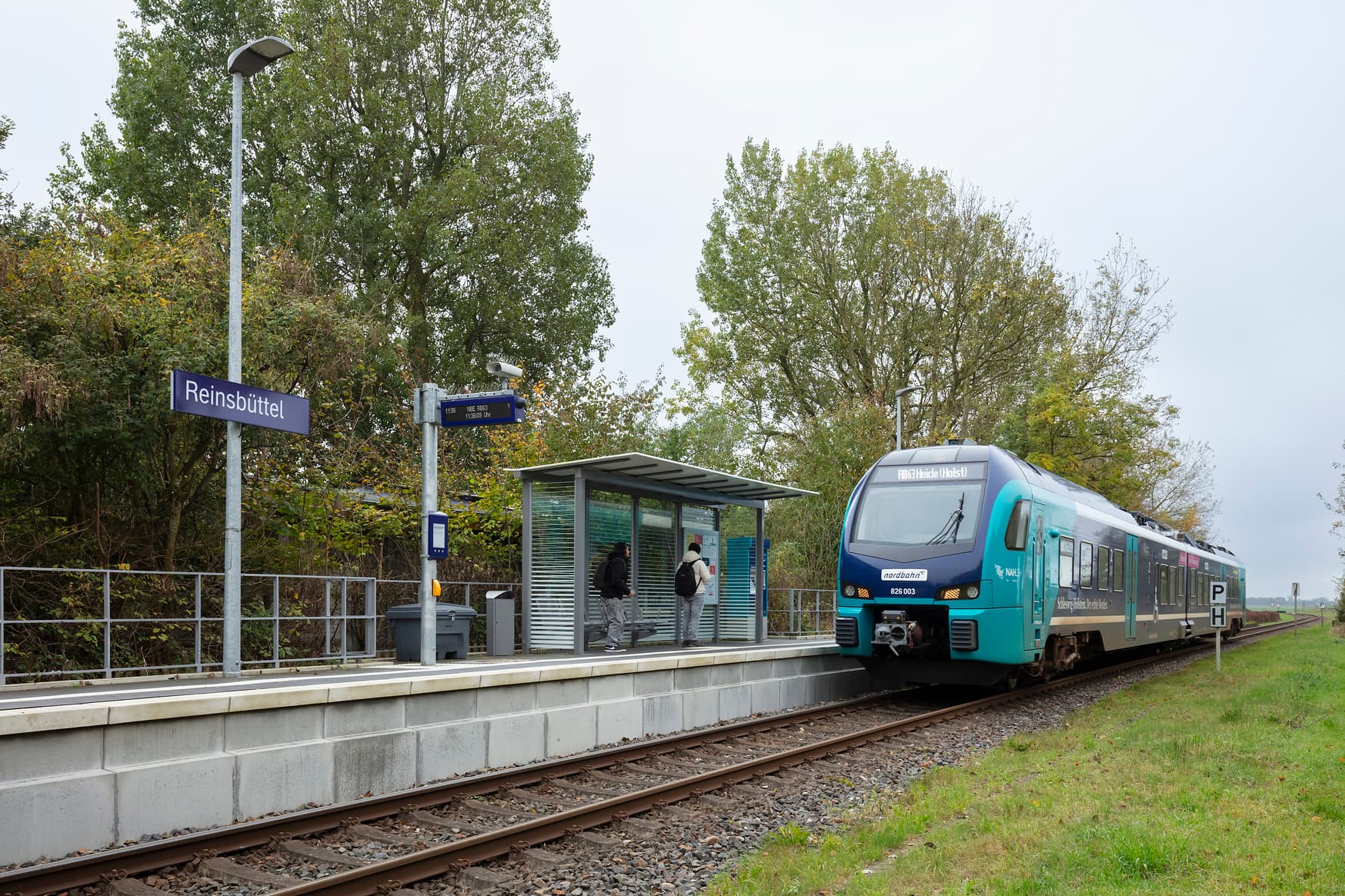 Neuer Bahnsteig mit Wetterschutzhäuschen und Digitaler Bauinfoanzeige. Im Wetterschutzhäuschen warten zwei Personen auf einen einfahrenden Zug.
