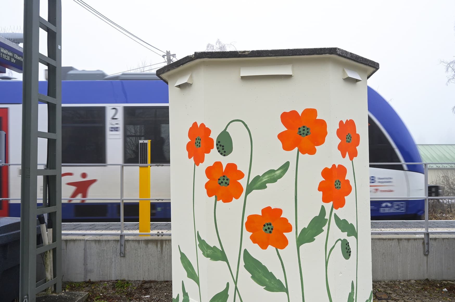 Schalthaus mit Klatschmohn bemalt, dahinter der Bahnsteig mit fahrendem Zug.