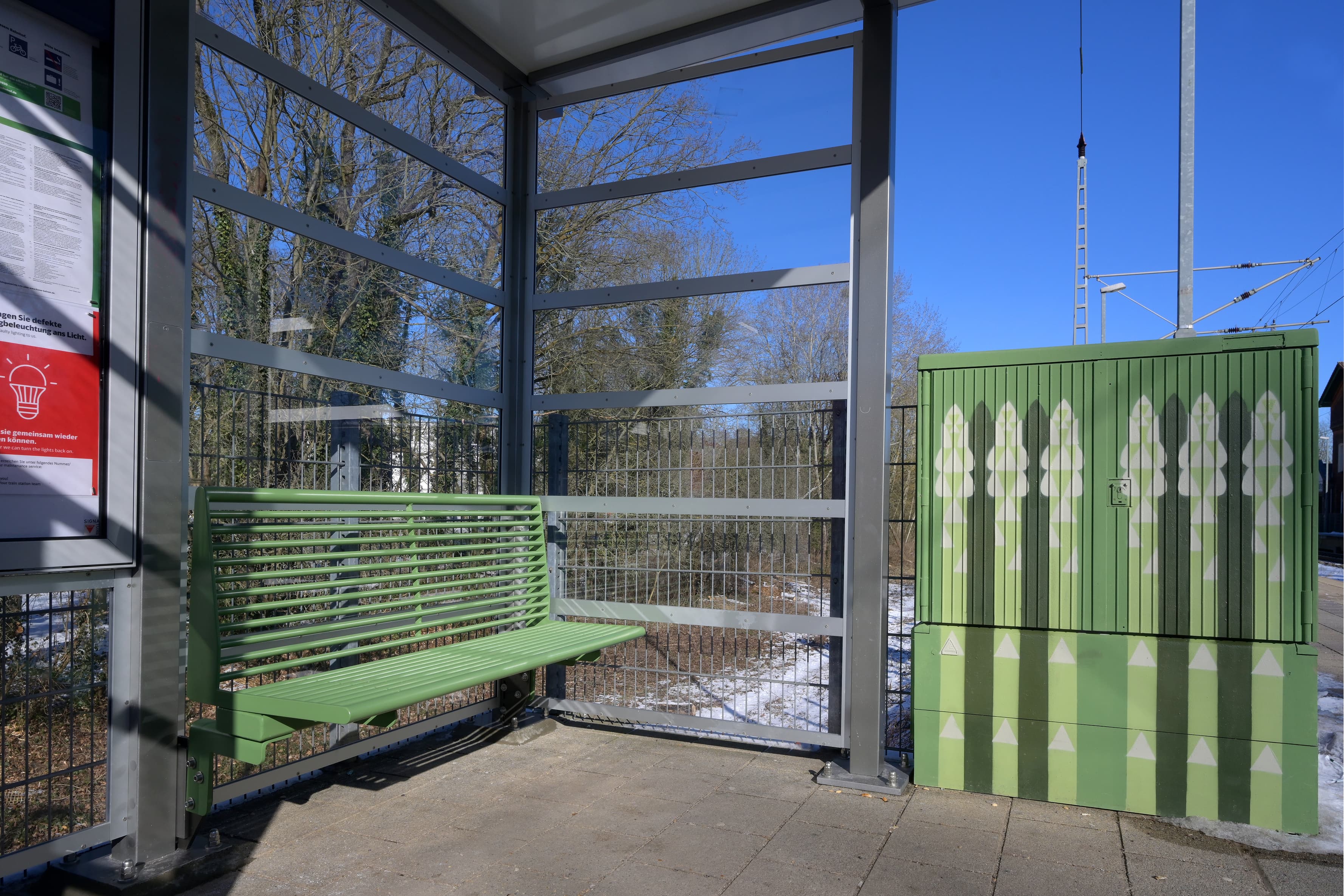 Eine grüne Bank in einem Wartehaus und rechts daneben ein grüner Stromkasten. Stromkasten