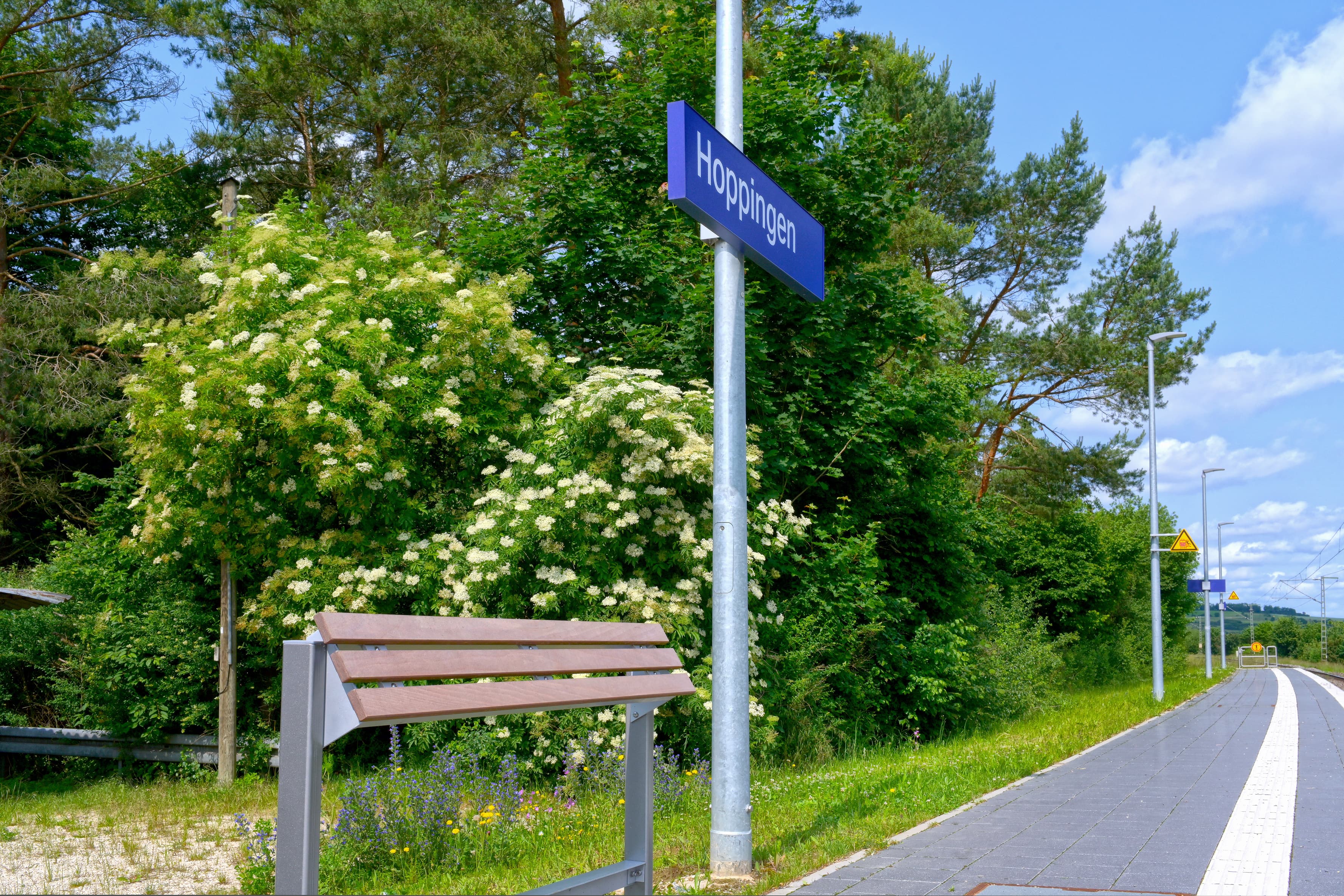 Lehnmöbel aus Holz und Metall links im Bild. Darüber Bahnhofsschild mit dem Bahnhofsnamen Hoppingen, rechts im Bild Gleise und ein gepflasterter Bahnsteig mit taktilem Leitsystem. 