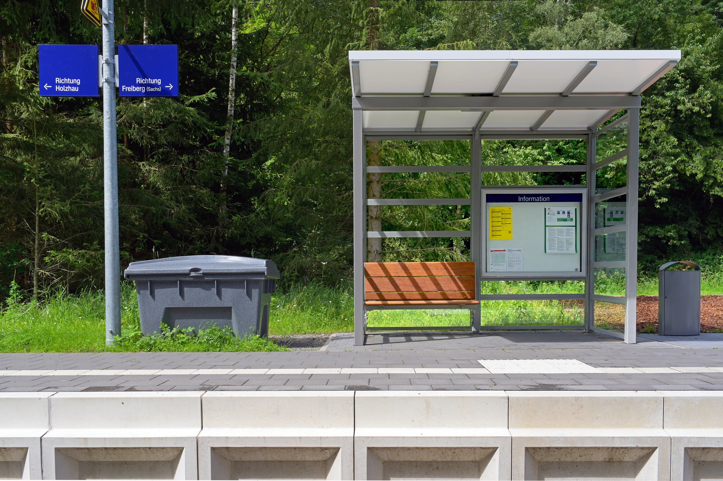 Zu sehen sind zwei Richtungsschilder an einem Bahnsteig, daneben ein grauer Streugutbehälter, rechts daneben direkt ein kleines, volltransparentes Wetterschutzhaus mit einer Vitrine und einer Holzbank mit Platz für bis zu 3 Personen. Am Bildrand sieht man dann auch noch eine grauen Mülleimer.