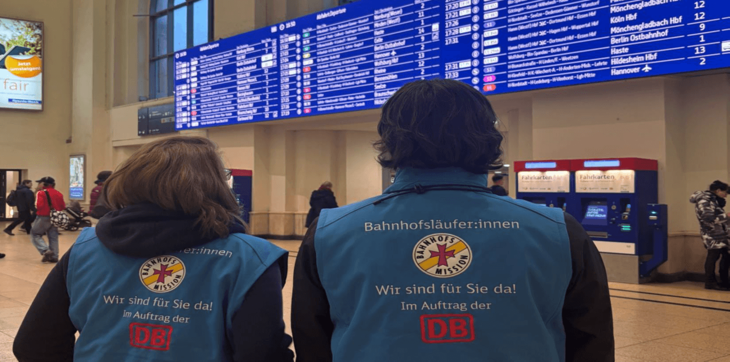 Die Rückenansicht von zwei Bahnhofsläufer:innen vor einer Abfahrtstafel in einem Bahnhof.