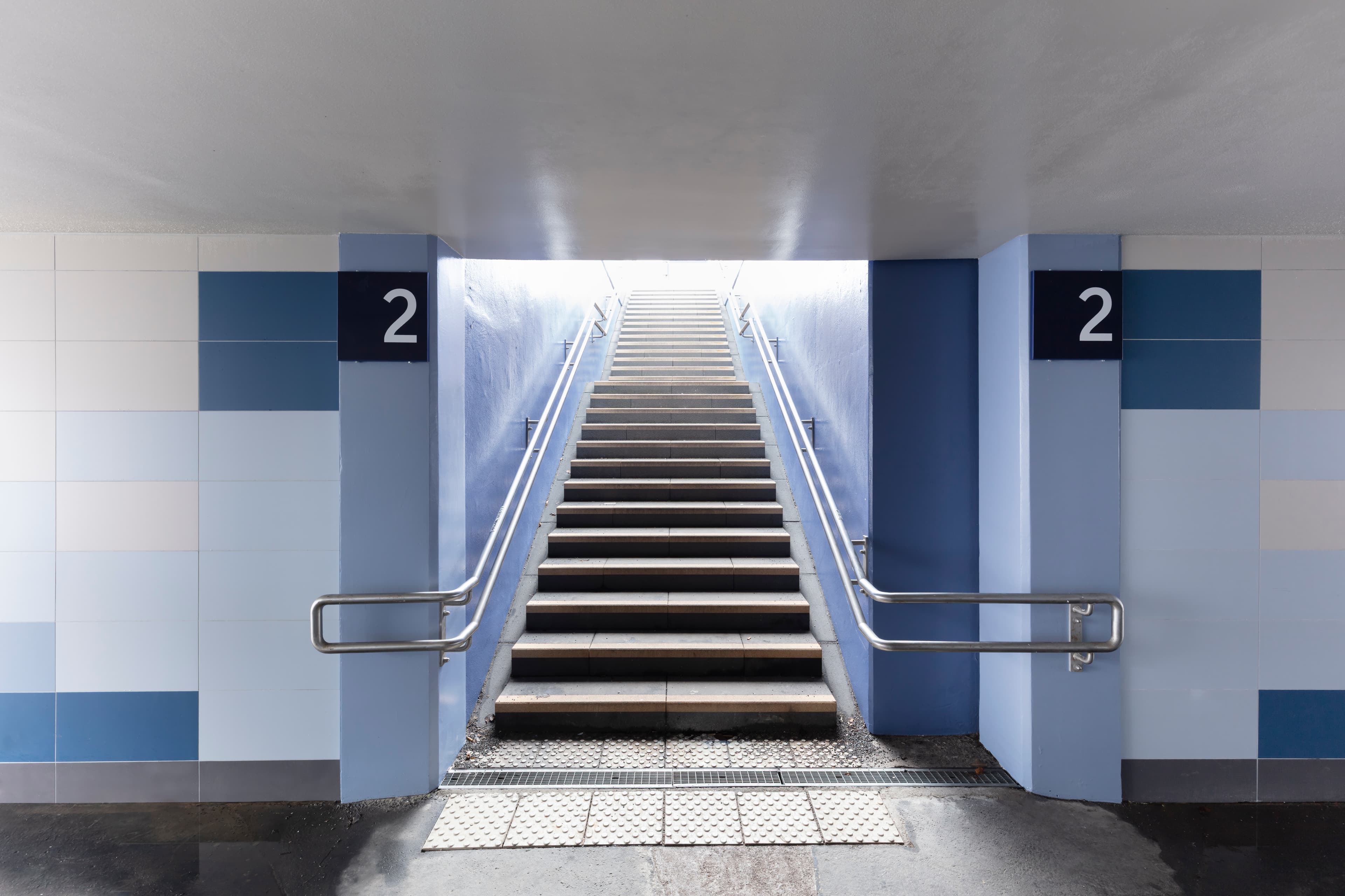 In der blau gefliesten Personenunterführung führt eine Treppe mit Handläufen auf beiden Seiten zum Bahnsteig 2.