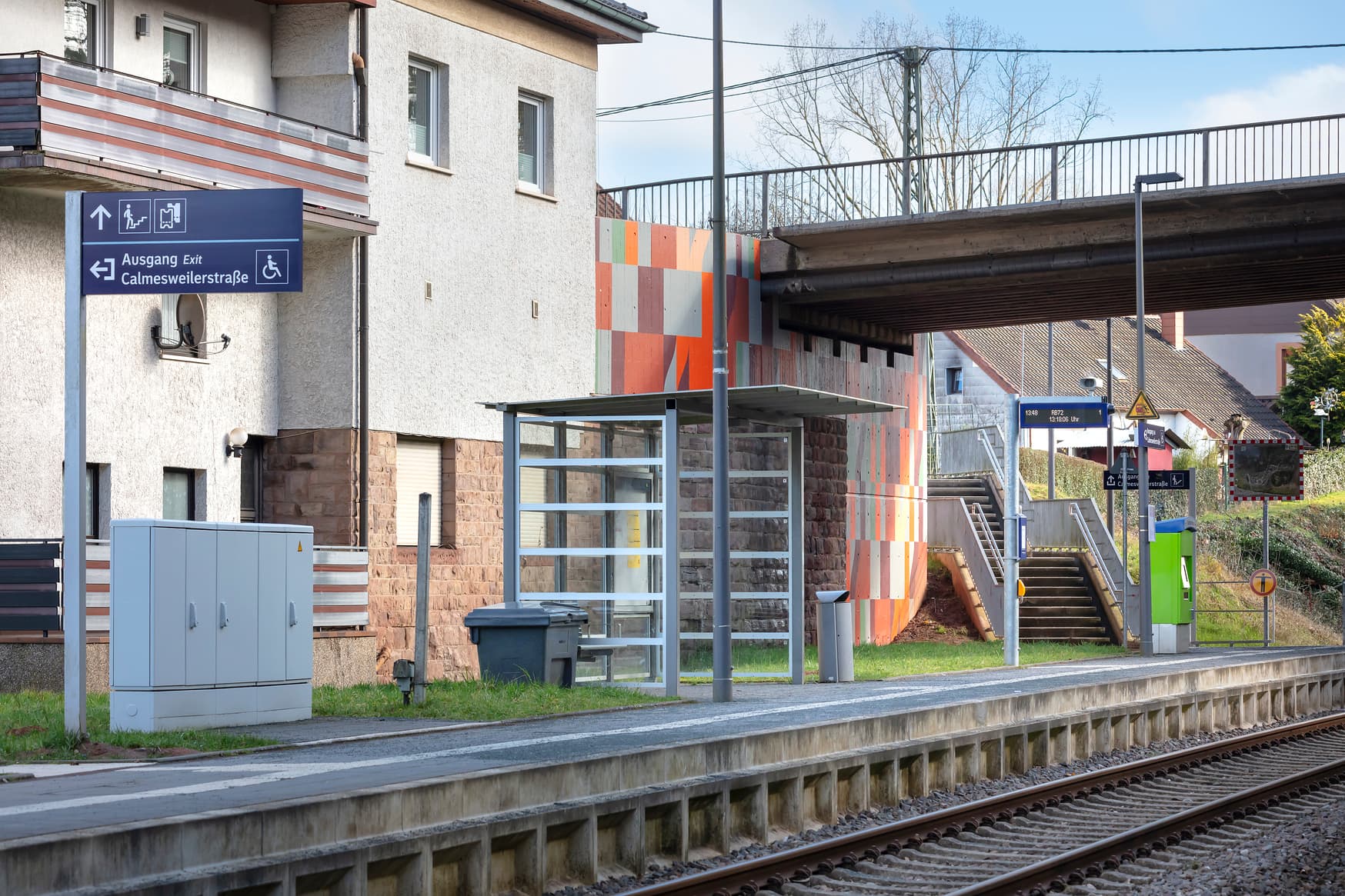 Bahnsteig mit Wetterschutzhaus, dynamischen Schriftenanzeiger, Beleuchtung, Treppenaufgang und bunt gestalteter Brückenpfeiler.