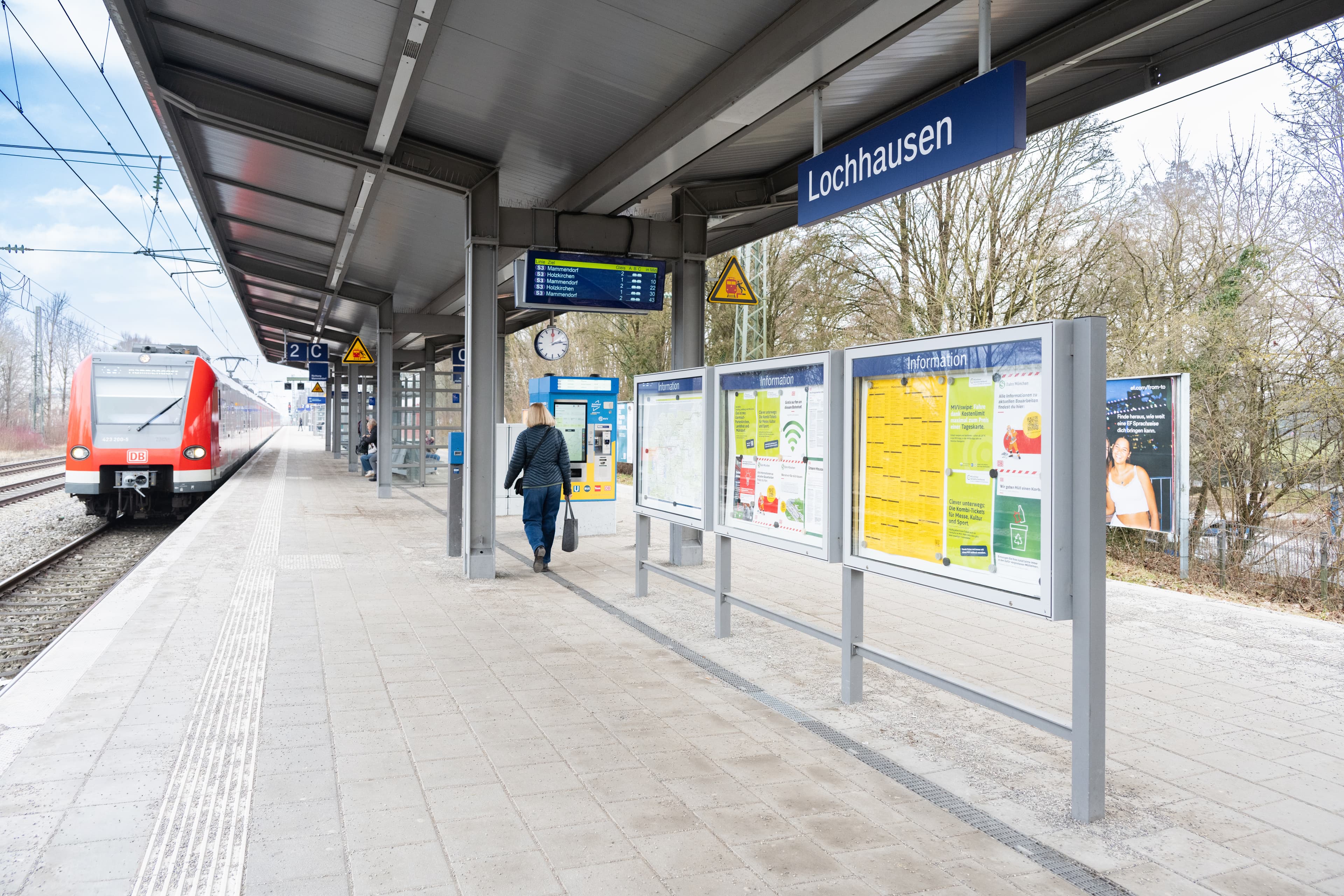 Links im Bild zu sehen ein einfahrender Zug der deutschen Bahn. Vordergründig und in der Mitte, der neue Bahnsetig des ZKB München Lochhausen mit Bahnhofsschild, Infovitrinen und taktilem Leitstreifen. 