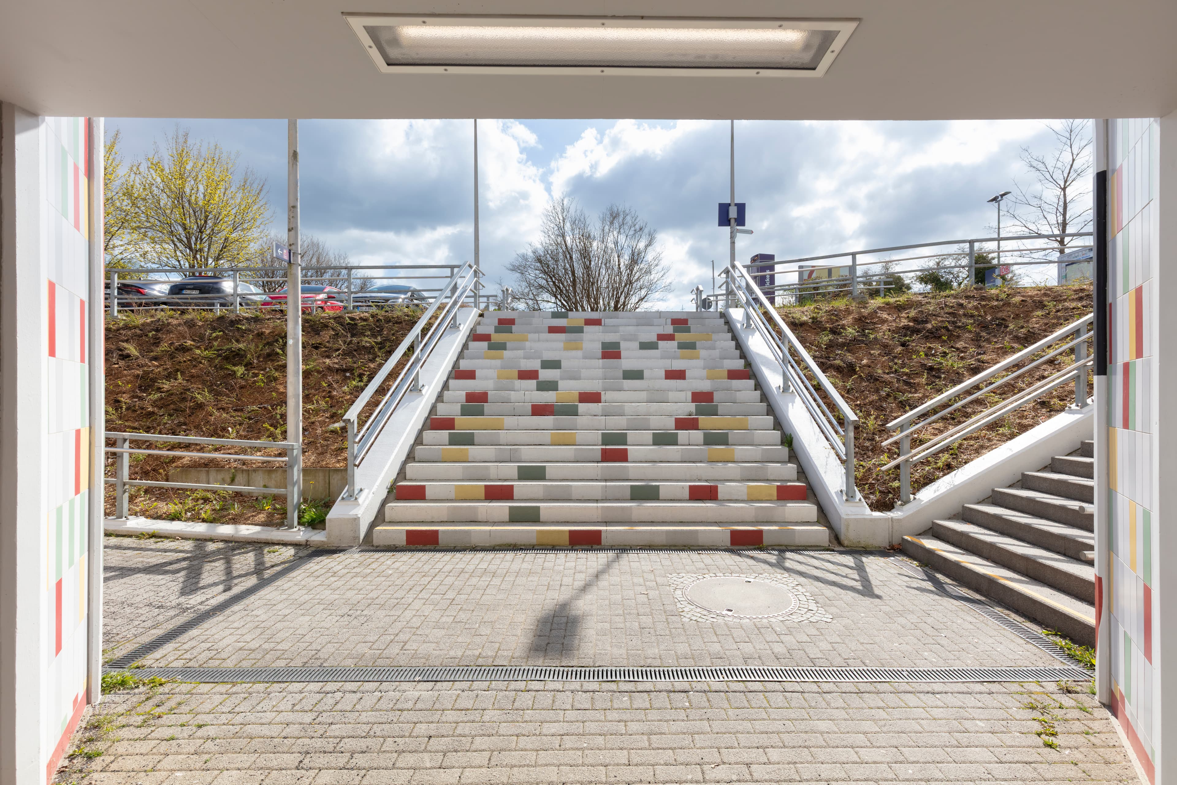 Geradeaus eine breite Treppe mit bunten Stufen. Rechts und links davon Bepflanzung.