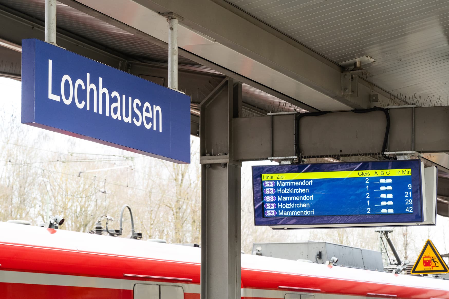 Blick auf Digitalen Zuganzeiger am Bahnhof Lochhausen (blaues Schild). Im Hintergrund ist das Dach von einem Zug zu erkennen. 