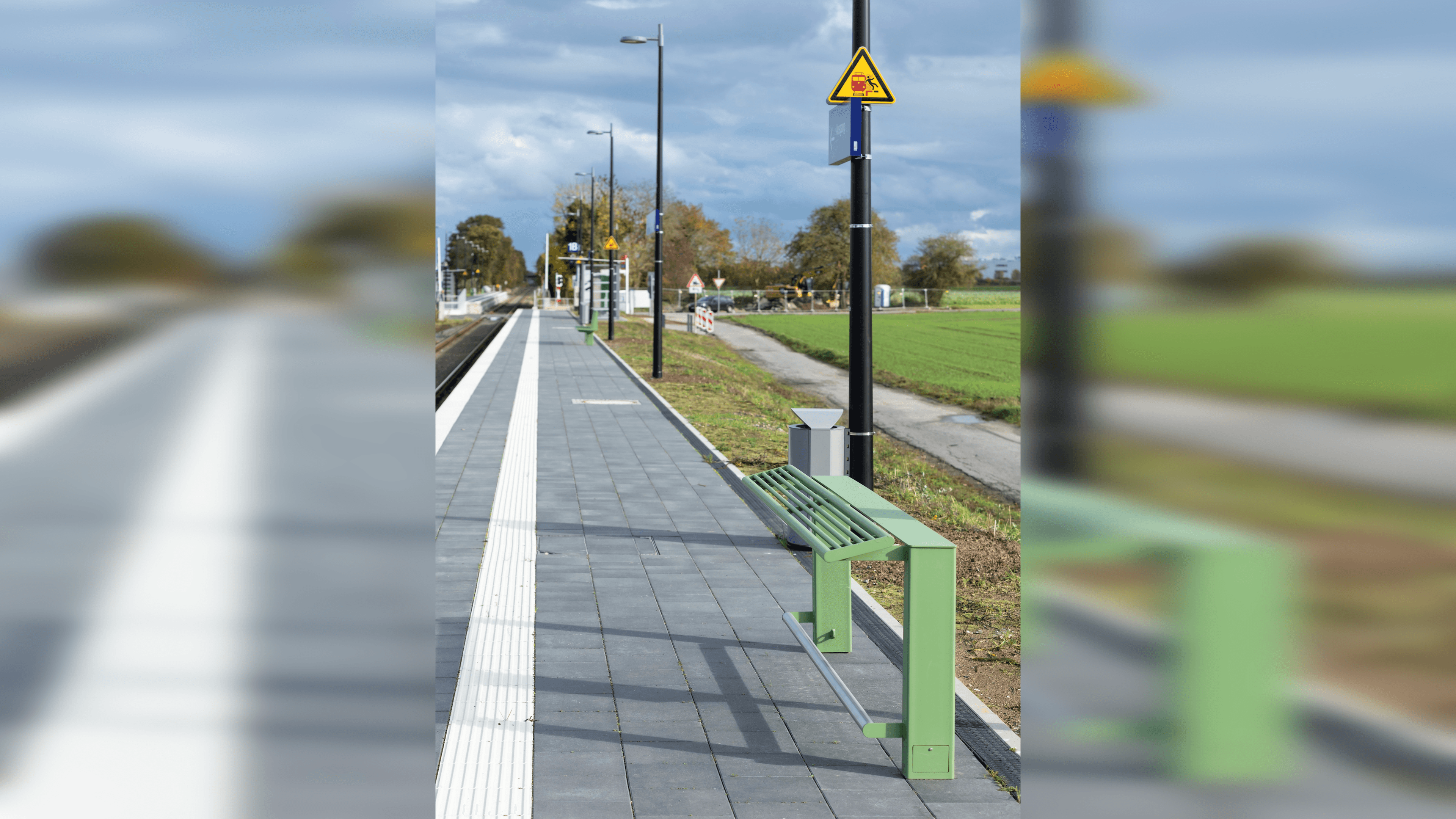 Bahnsteig mit taktilem Leitsystem. Rechts befindet sich ein grüner Anlehner. Im Hintergrund sind Laternen und ein Wetterschutzhäuschen zu sehen. Rechts vom Bahnsteig ist eine grüne Wiese und links das Gleis.