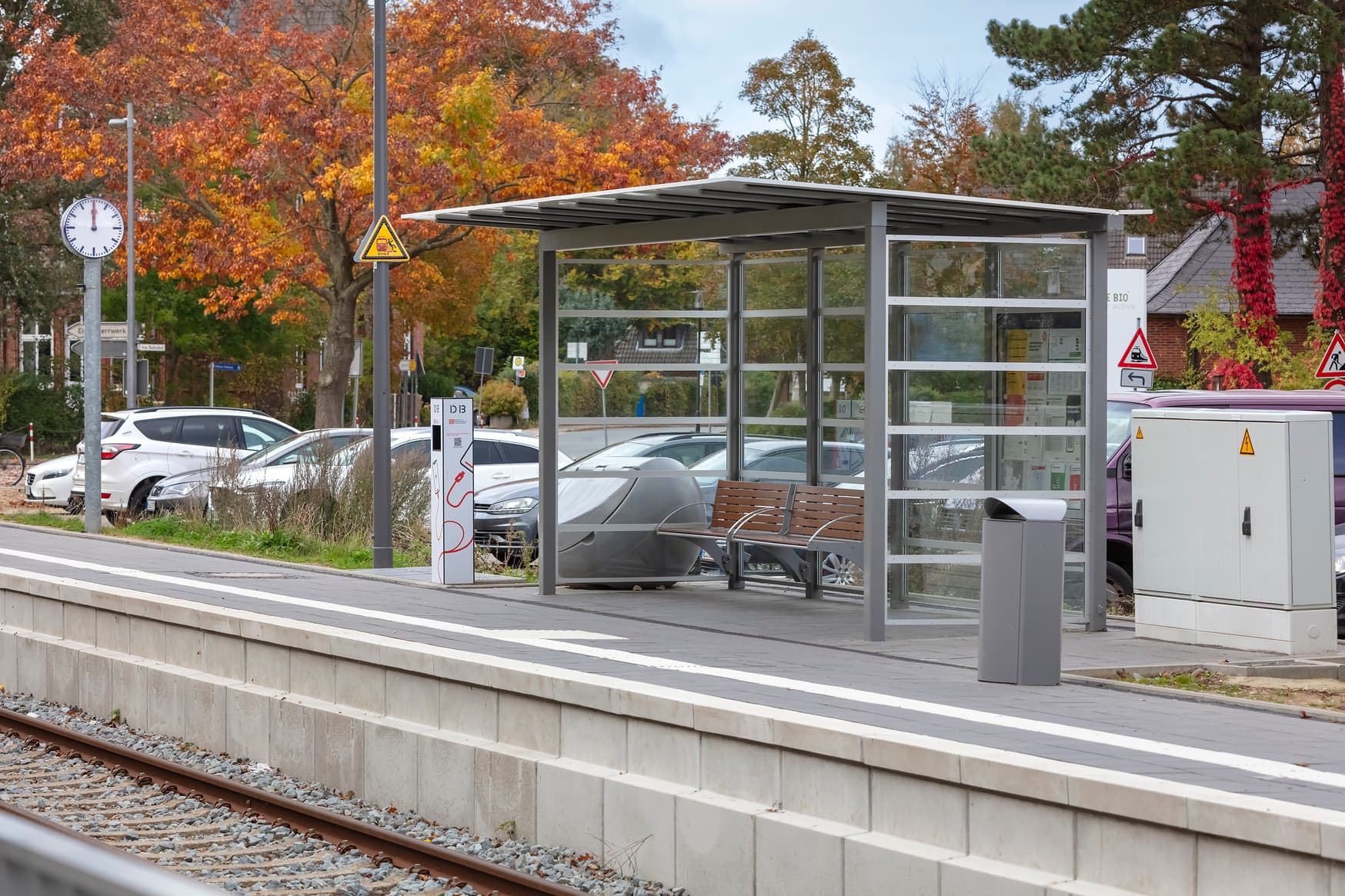 Wetterschutzhäuschen mit Sitzbank aus Holz daneben Solarbetriebene Handyladesäule, weiter hinten auf dem Bahnsteig die Bahnhofsuhr.