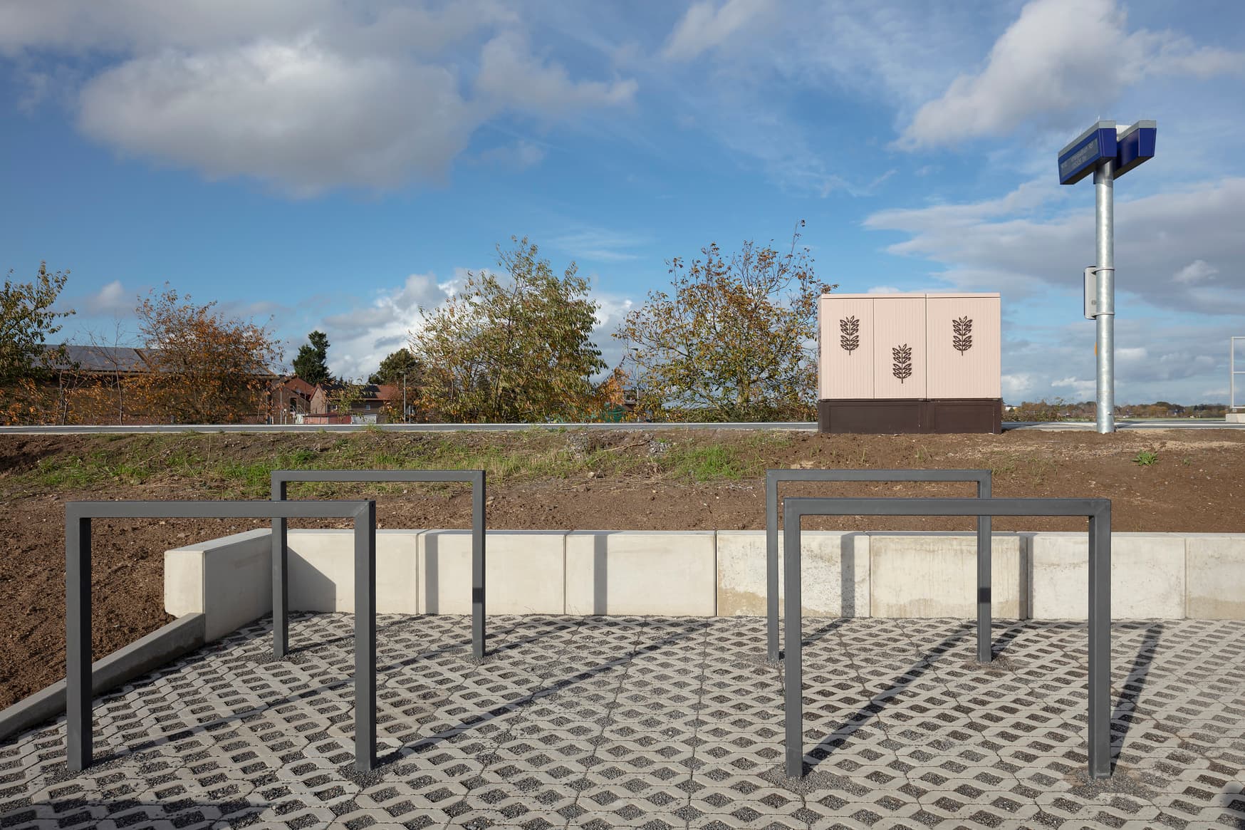 Vier Fahrradbügel in dunkelgrauer Farbe in einer gepflasterten Einbruchtung neben dem Bahnsteig. Das Technikhäuschen und der Dynamische Schriftenanzeiger sind auf dem Bahnsteig.